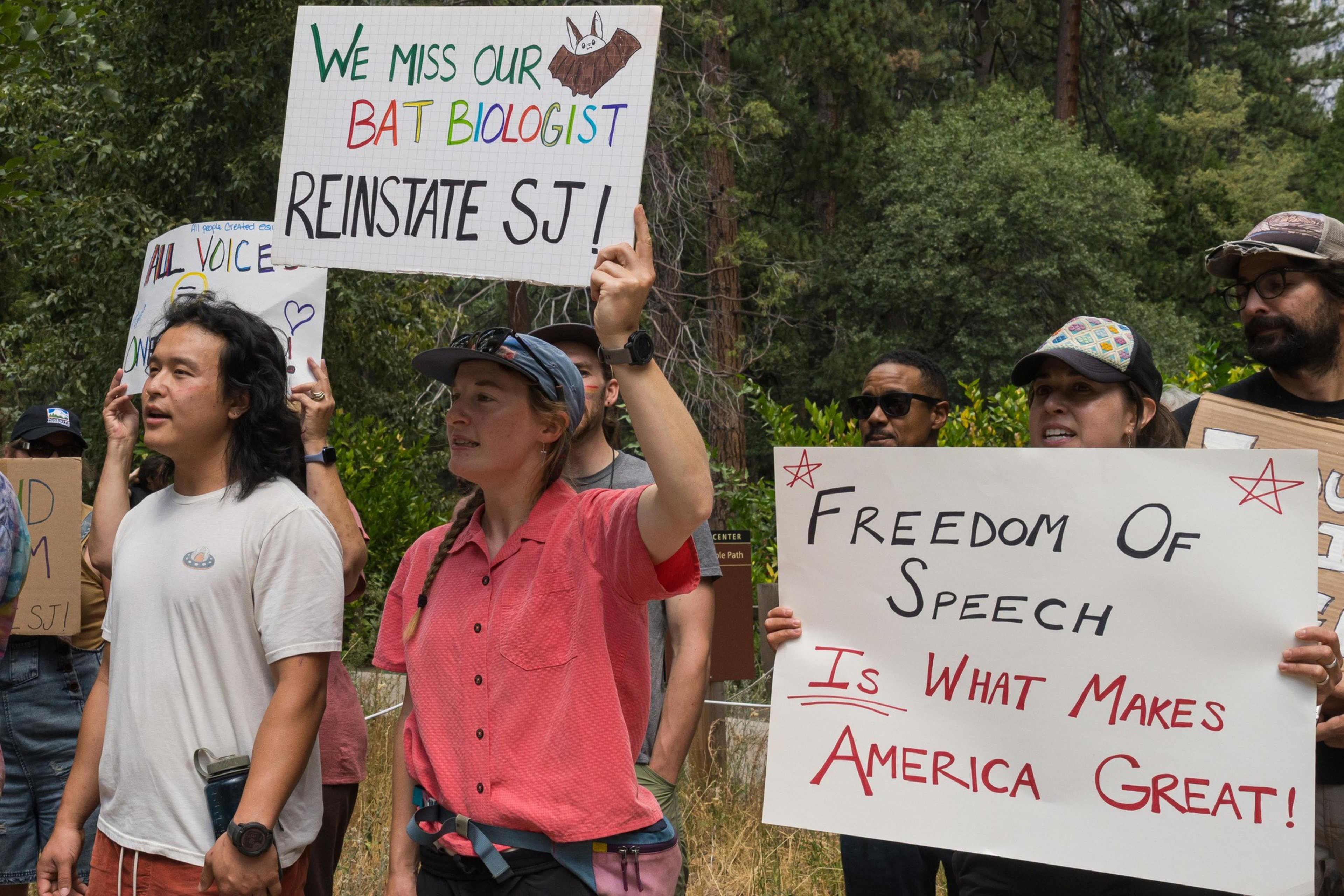People hold signs reading "We miss our bat biologist, reinstate SJ!" and "Freedom of speech is what makes America great!" during an outdoor protest.