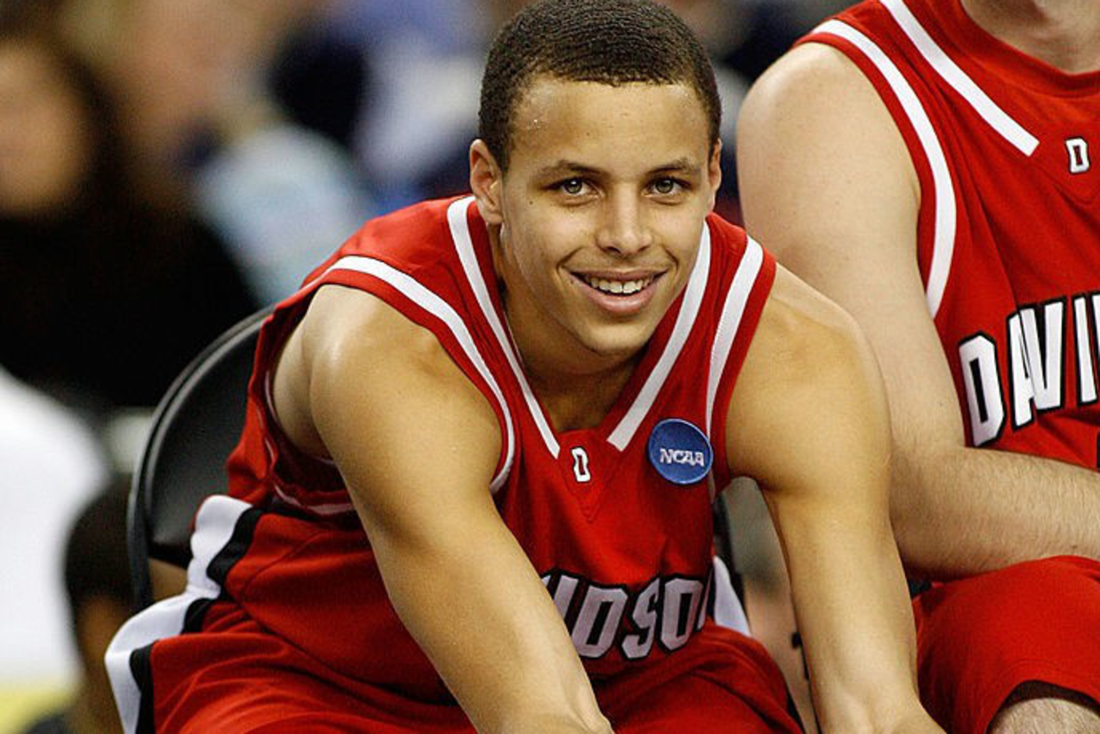A basketball player in a red Davidson uniform is sitting down, stretching forward to touch his shoes, smiling, with a crowd blurred in the background.