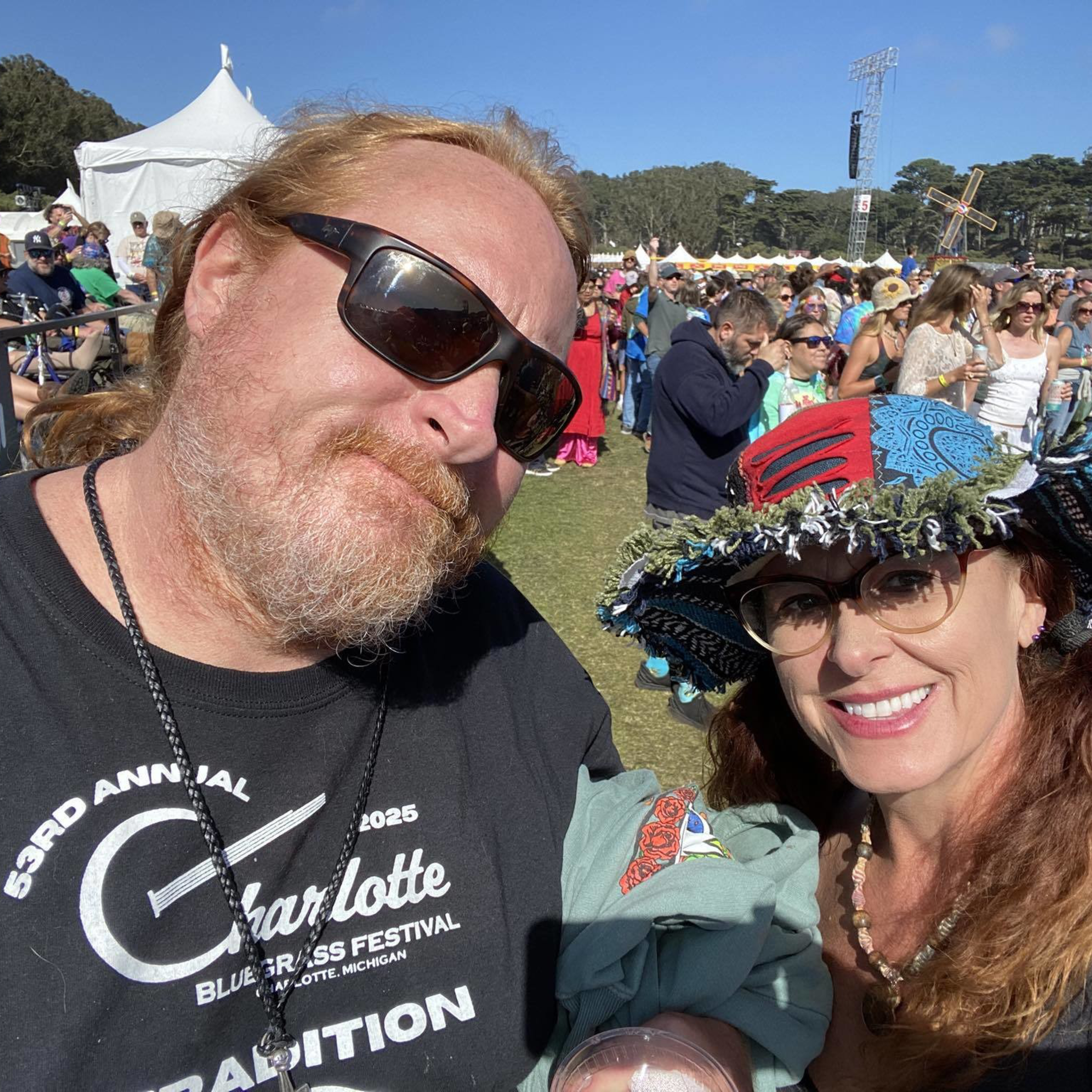 A man with sunglasses and a woman wearing a colorful hat smile at a sunny outdoor festival with a crowd and tents in the background.