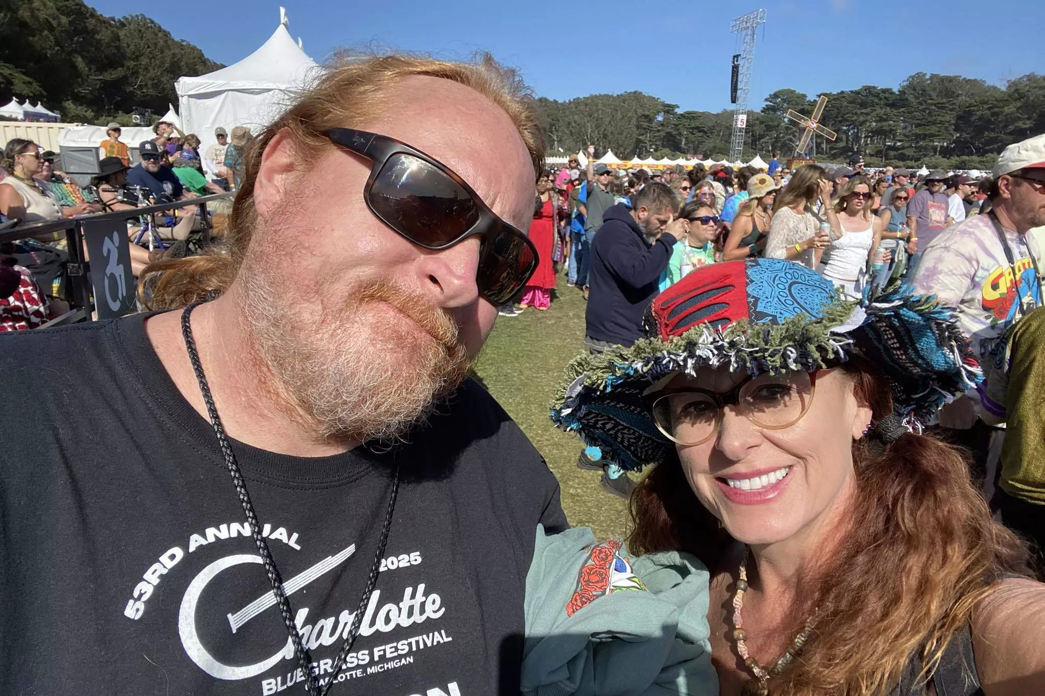A man with sunglasses and a woman wearing a colorful hat smile at a sunny outdoor festival with a crowd and tents in the background.