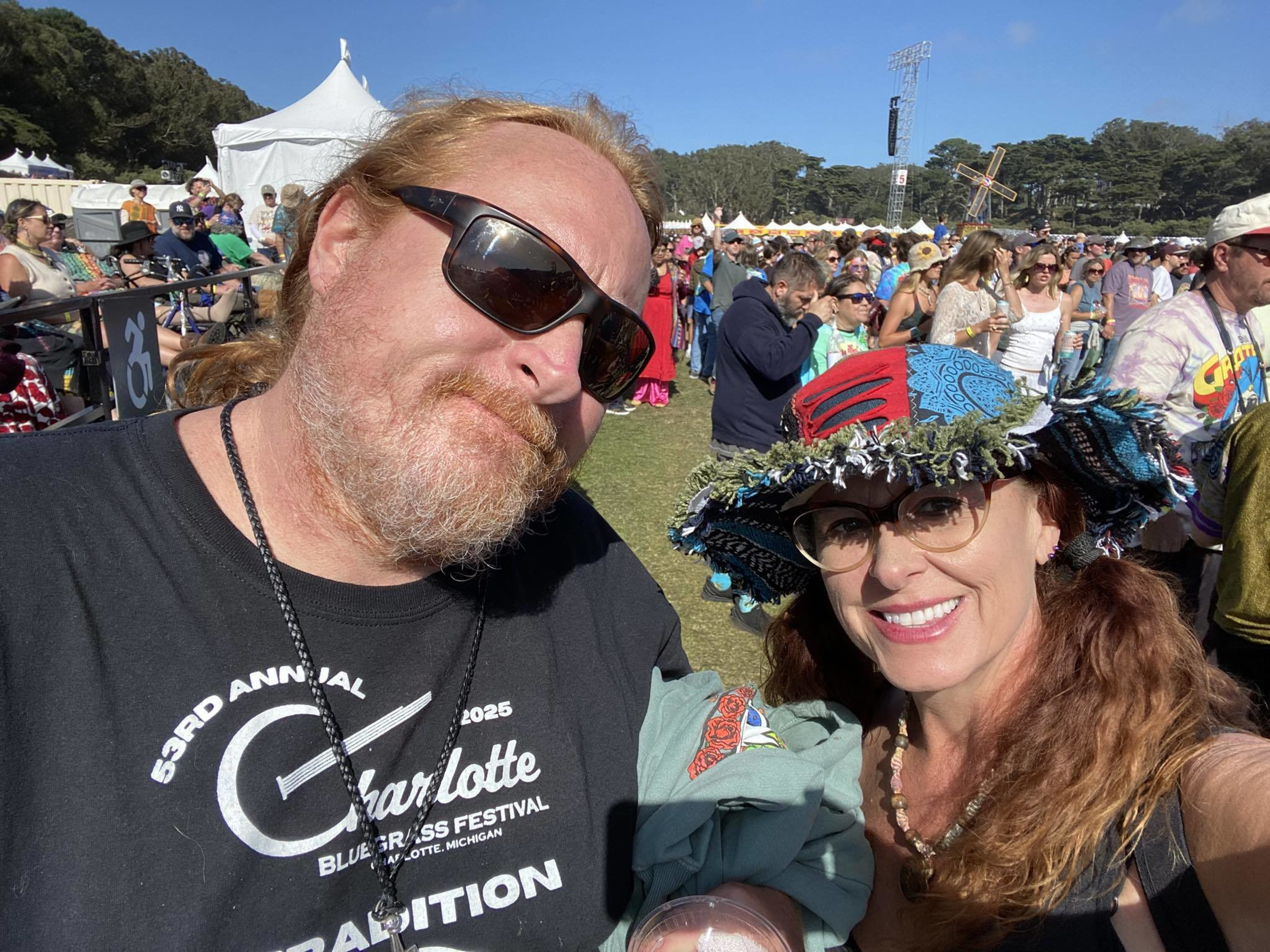 A man with sunglasses and a woman wearing a colorful hat smile at a sunny outdoor festival with a crowd and tents in the background.