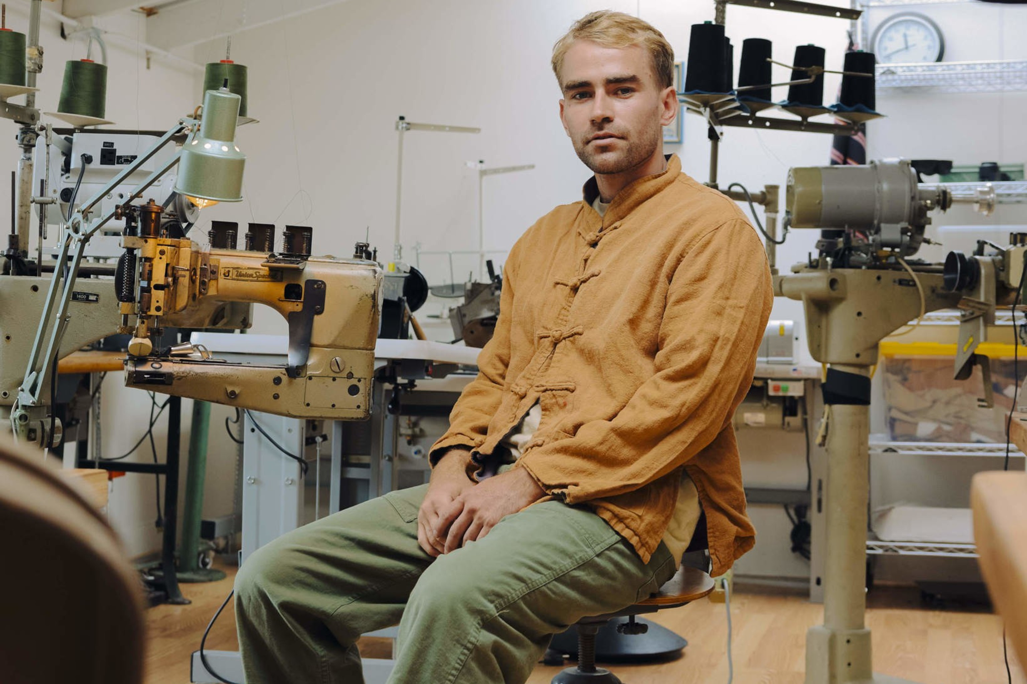 A man in a mustard yellow shirt and green pants sits on a stool in a sewing workshop surrounded by industrial sewing machines.