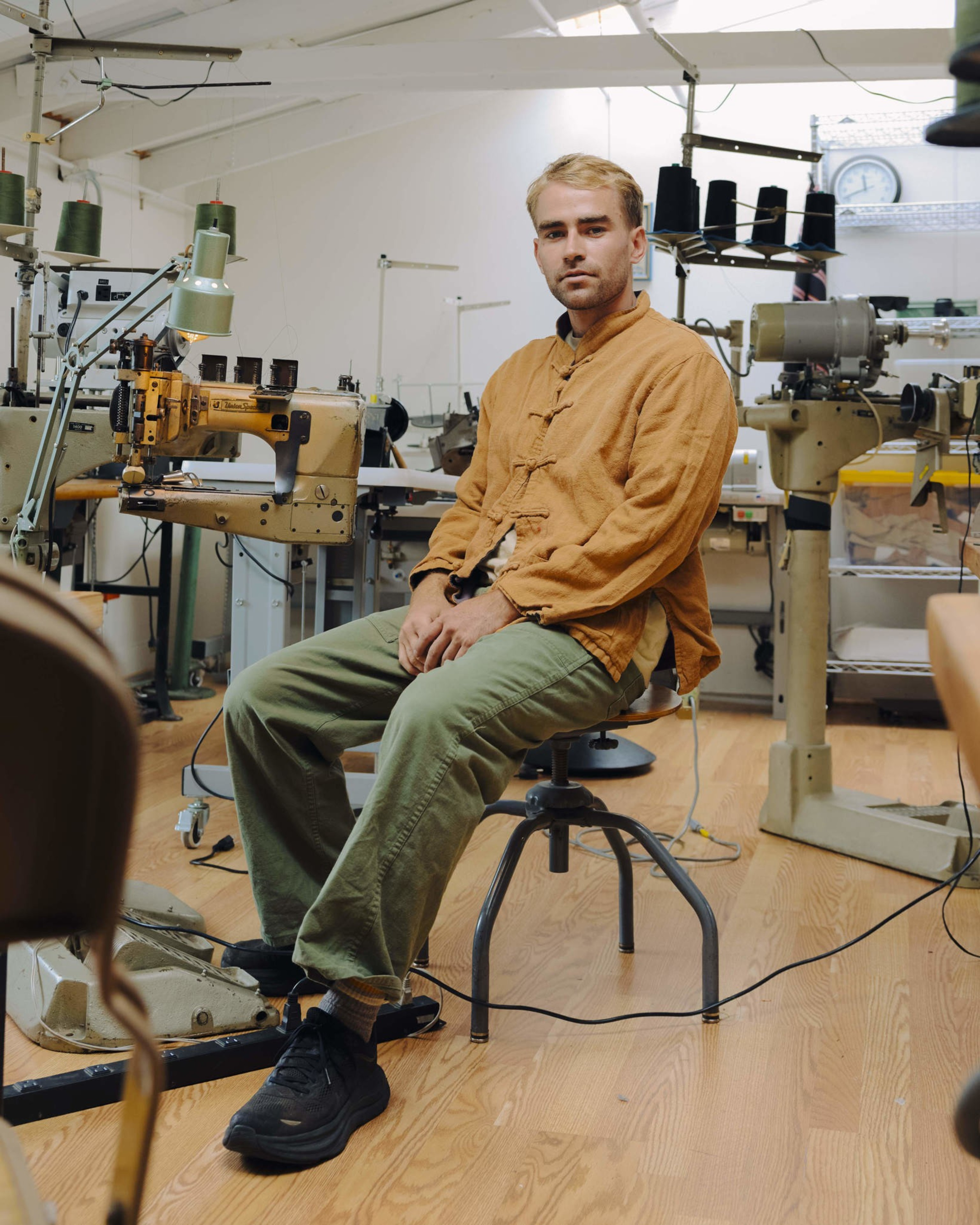 A man in a mustard yellow shirt and green pants sits on a stool in a sewing workshop surrounded by industrial sewing machines.