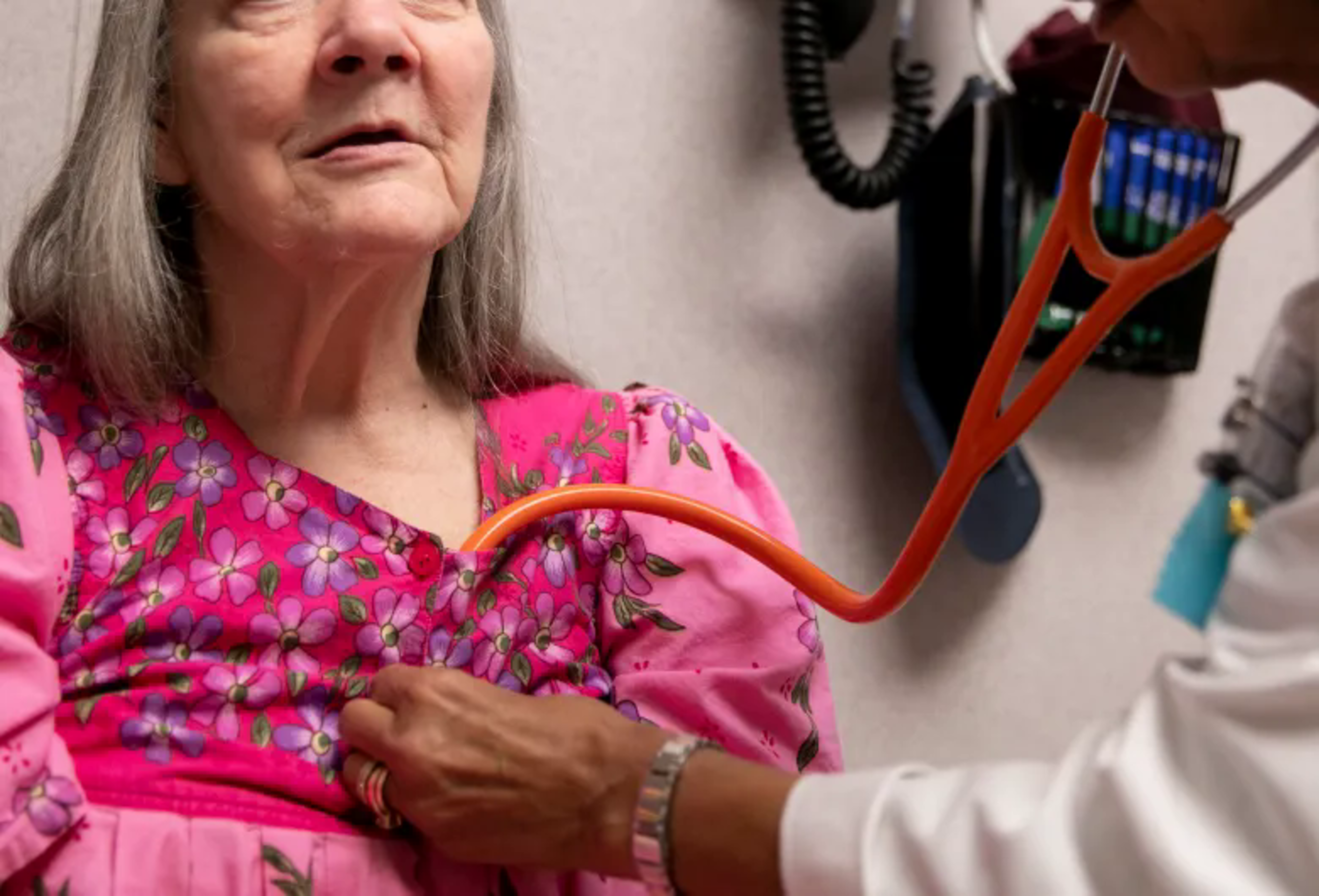 An elderly woman in a pink floral dress is being examined by a doctor using an orange stethoscope on her chest.