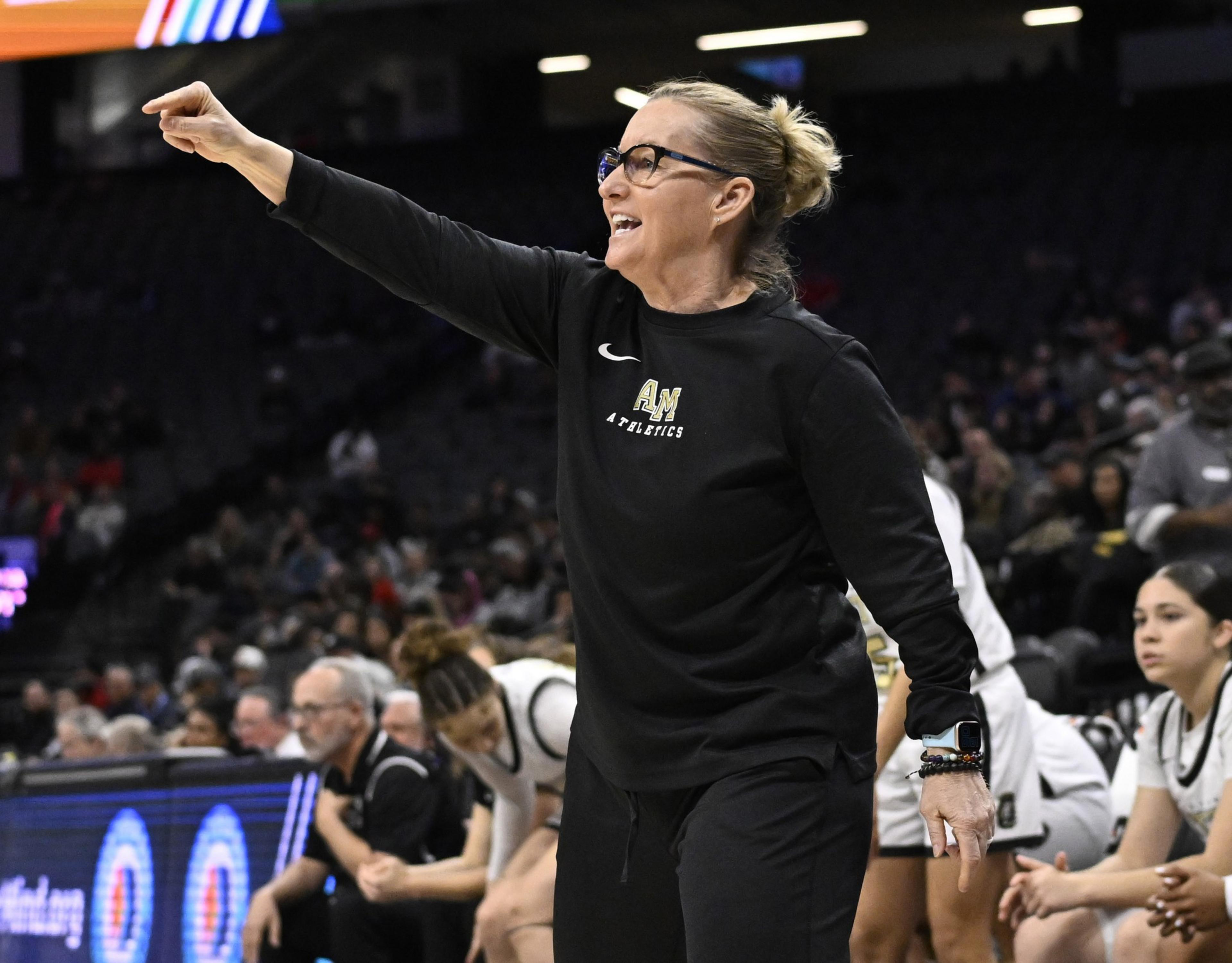 A coach enthusiastically gestures from the sidelines of a basketball game. She's wearing glasses and a black tracksuit, with players and spectators in the background.
