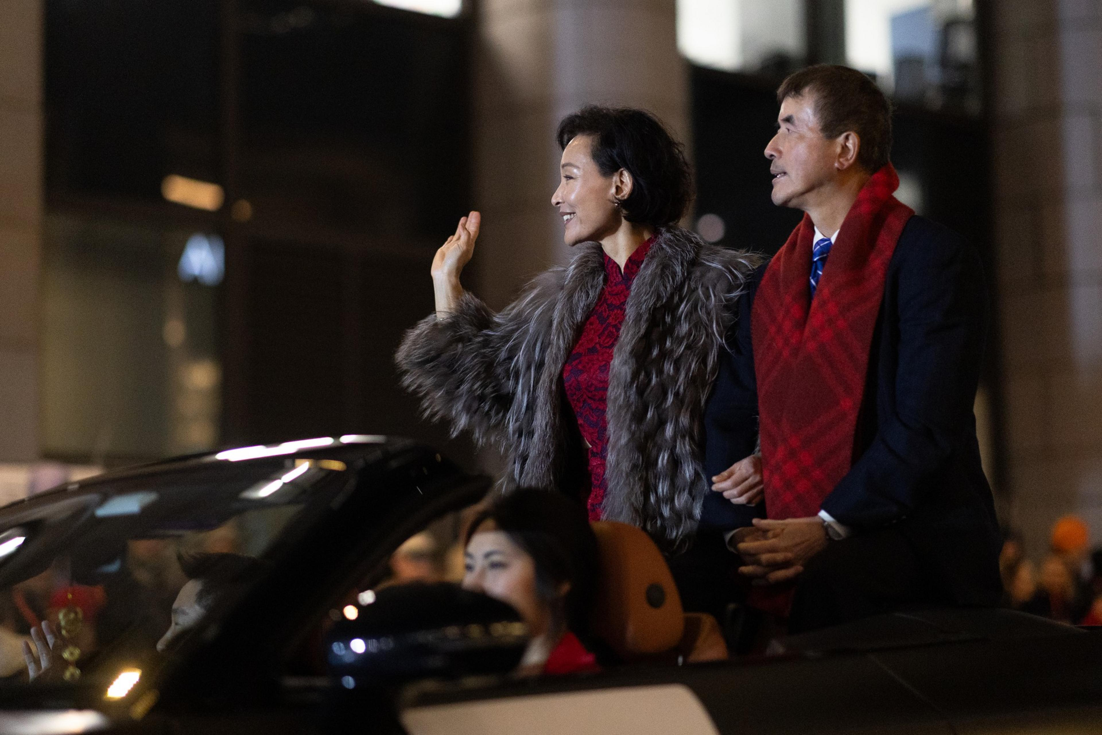 A woman in a fur coat waves while standing next to a man in a suit and red scarf, both seated in a convertible with a driver in front.