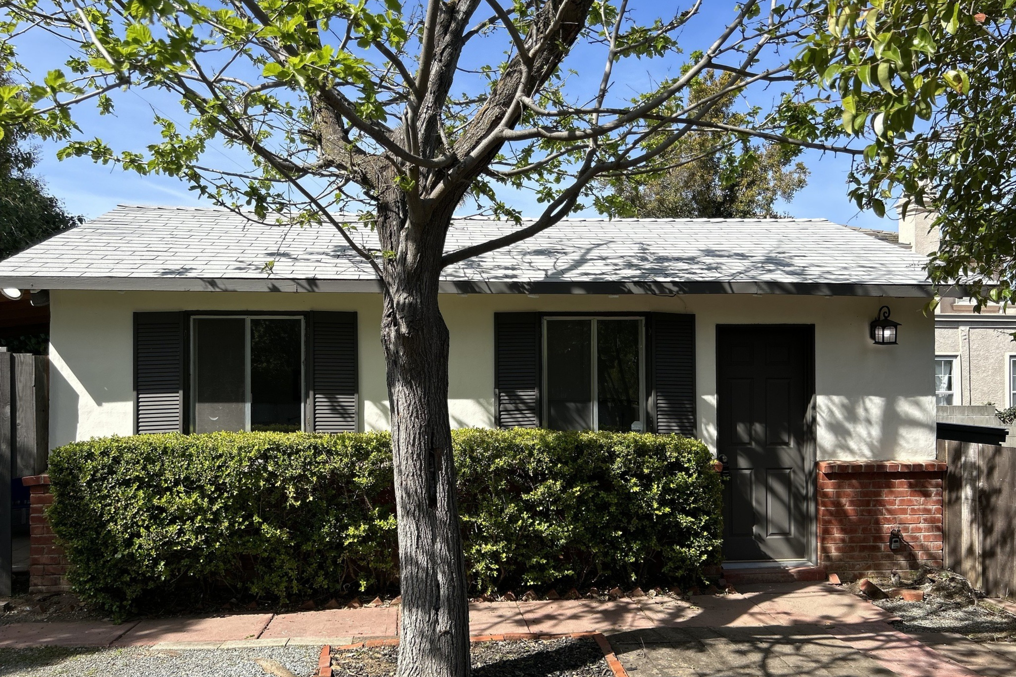 A single-story house with a grey roof, white walls, black shutters, and a front hedge.