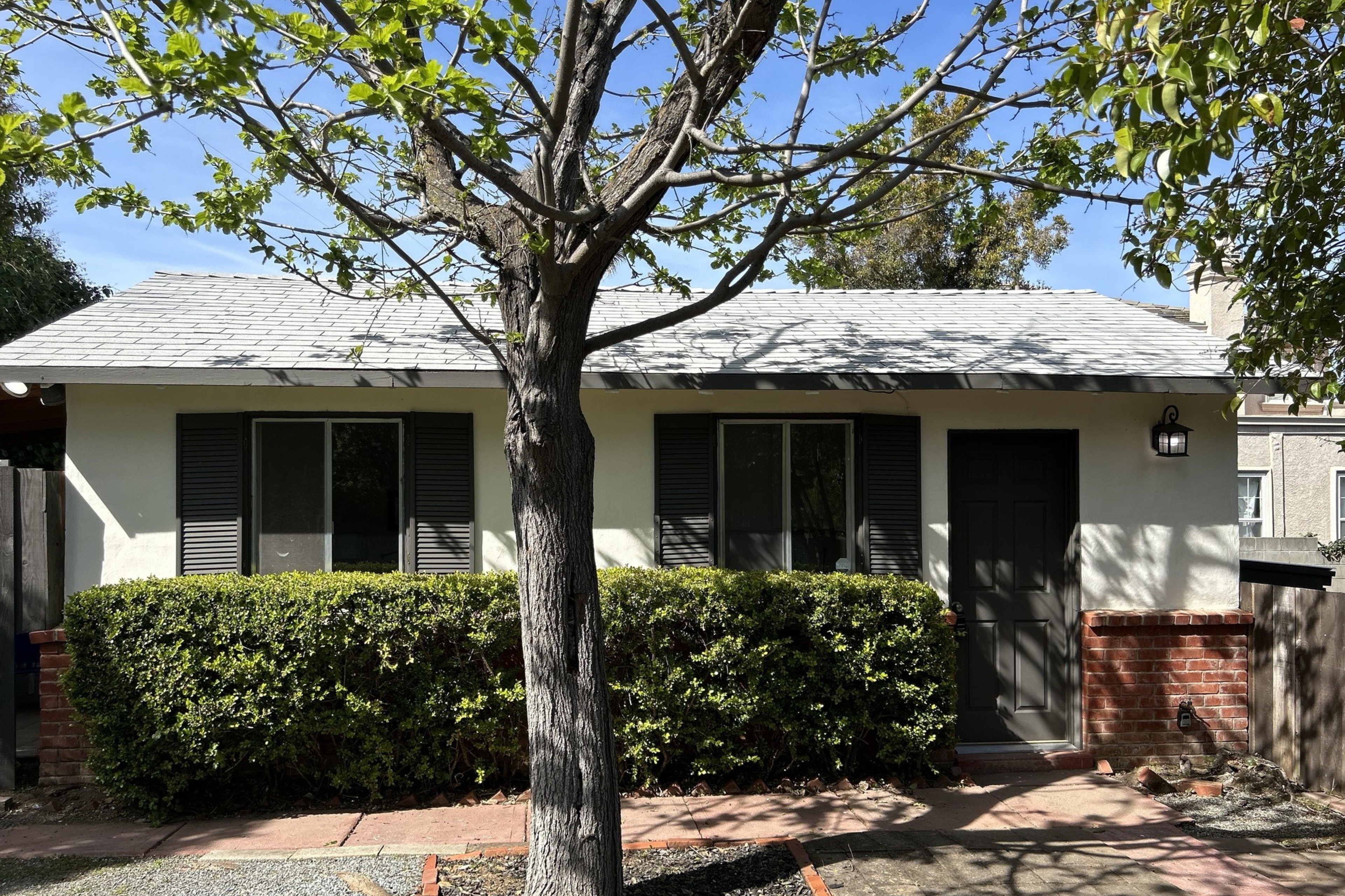 A single-story house with a grey roof, white walls, black shutters, and a front hedge.