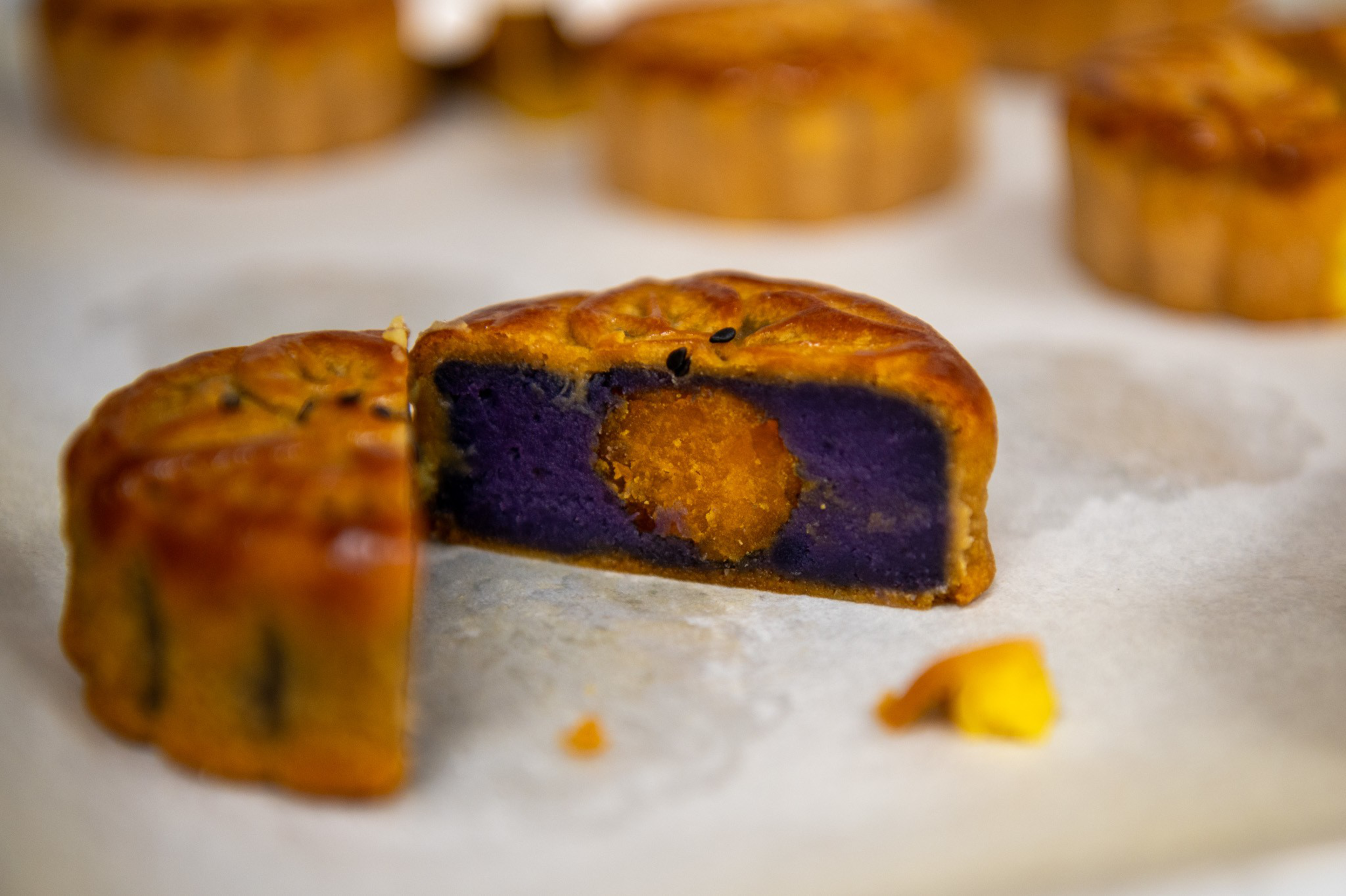 A golden-brown mooncake is cut in half to reveal a vivid purple filling with an orange yolk-like center, placed on a white surface. Several whole mooncakes are blurred in the background.