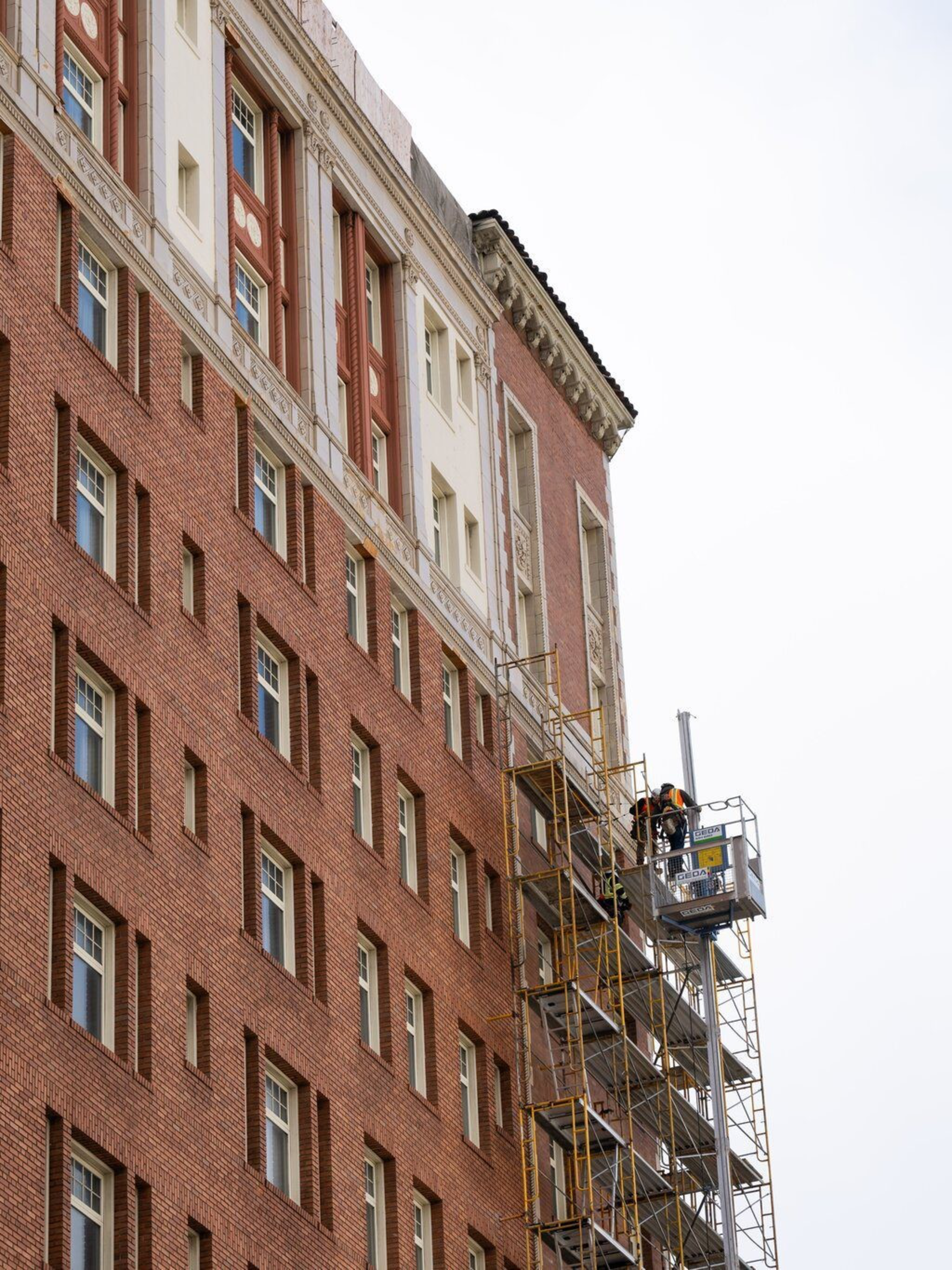 A brick building with symmetrical windows has fire escape stairs on the left side and scaffolding with workers on the right, against a partly cloudy sky.