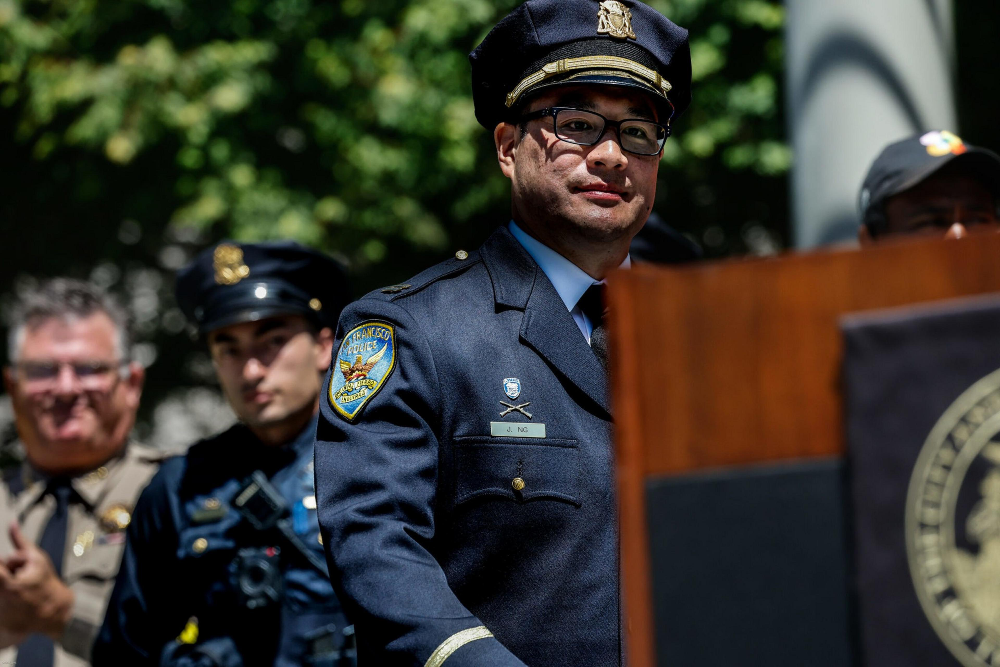 A police officer in uniform stands at a podium with other uniformed officers blurred in the background, set against a leafy outdoor backdrop.