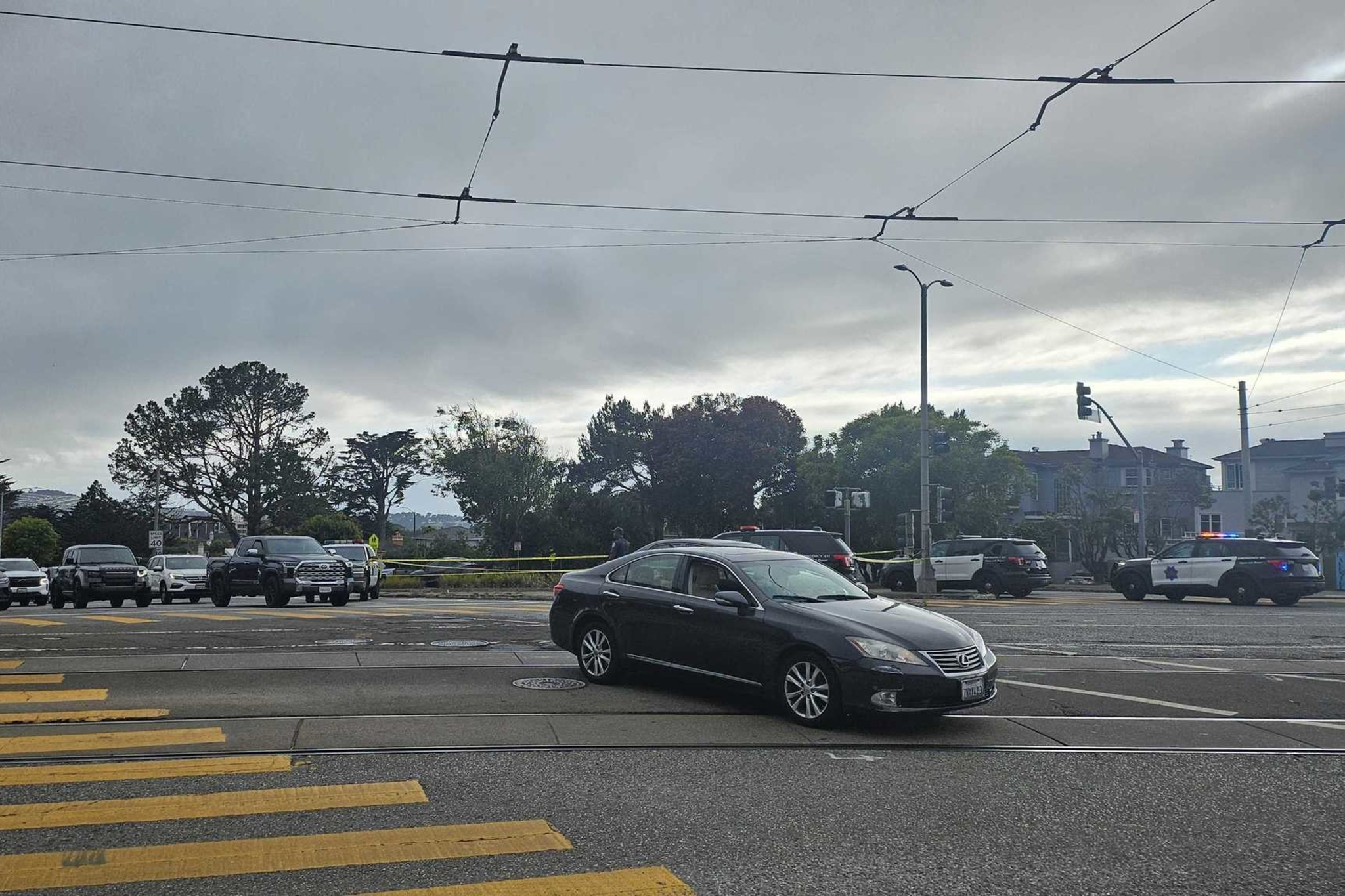 A black car is stopped diagonally in the middle of an intersection with several police vehicles and yellow caution tape blocking the surrounding area.