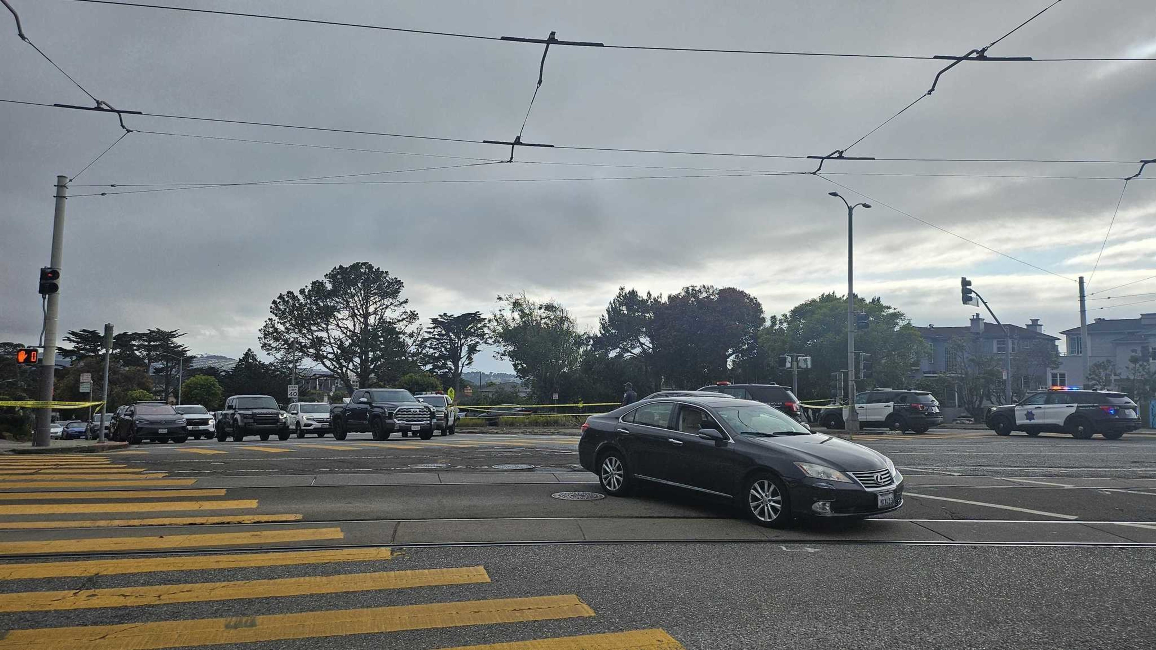 A black car is stopped diagonally in the middle of an intersection with several police vehicles and yellow caution tape blocking the surrounding area.