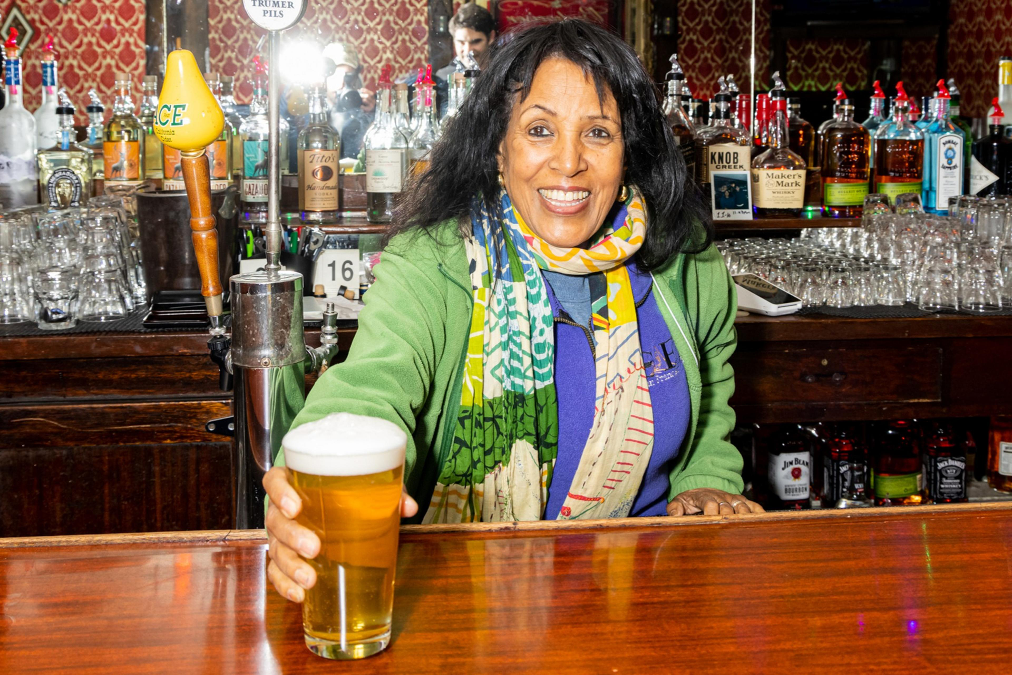 A smiling woman in a colorful scarf and green jacket stands behind a bar, offering a pint of beer. Bottles and glasses neatly line the shelves behind her.