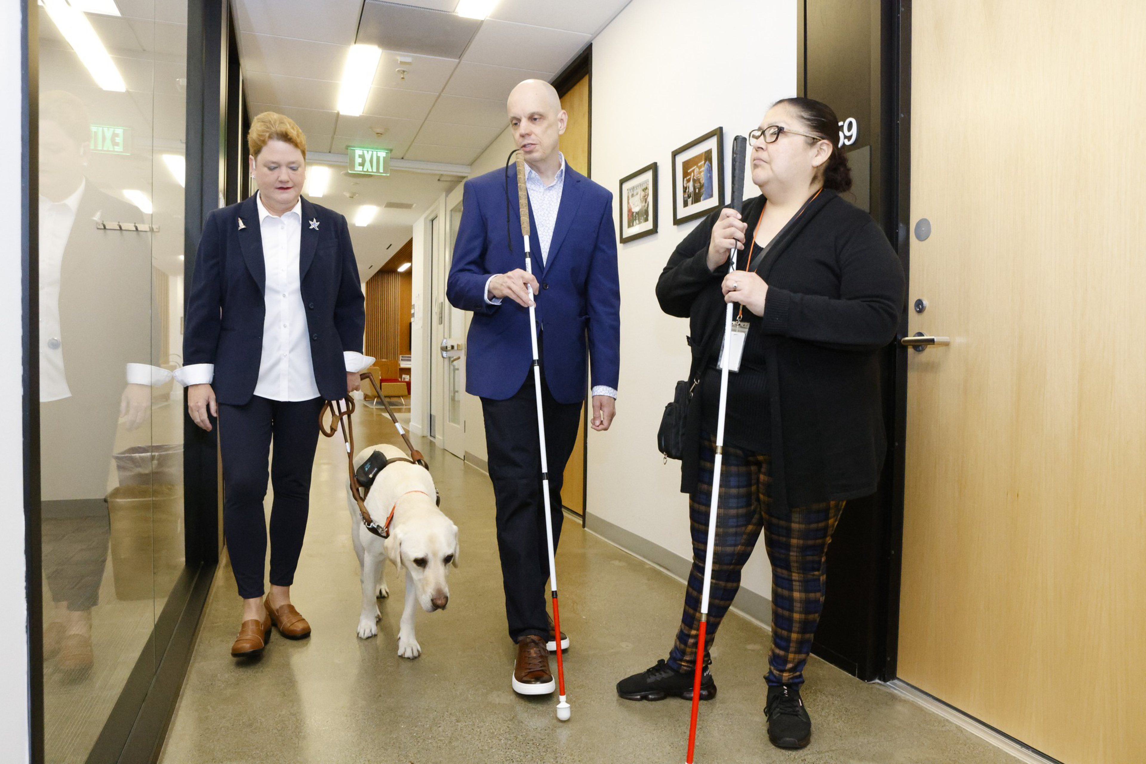 Three people are walking through a hallway, two holding white canes, one with a guide dog, while the third person accompanies them.