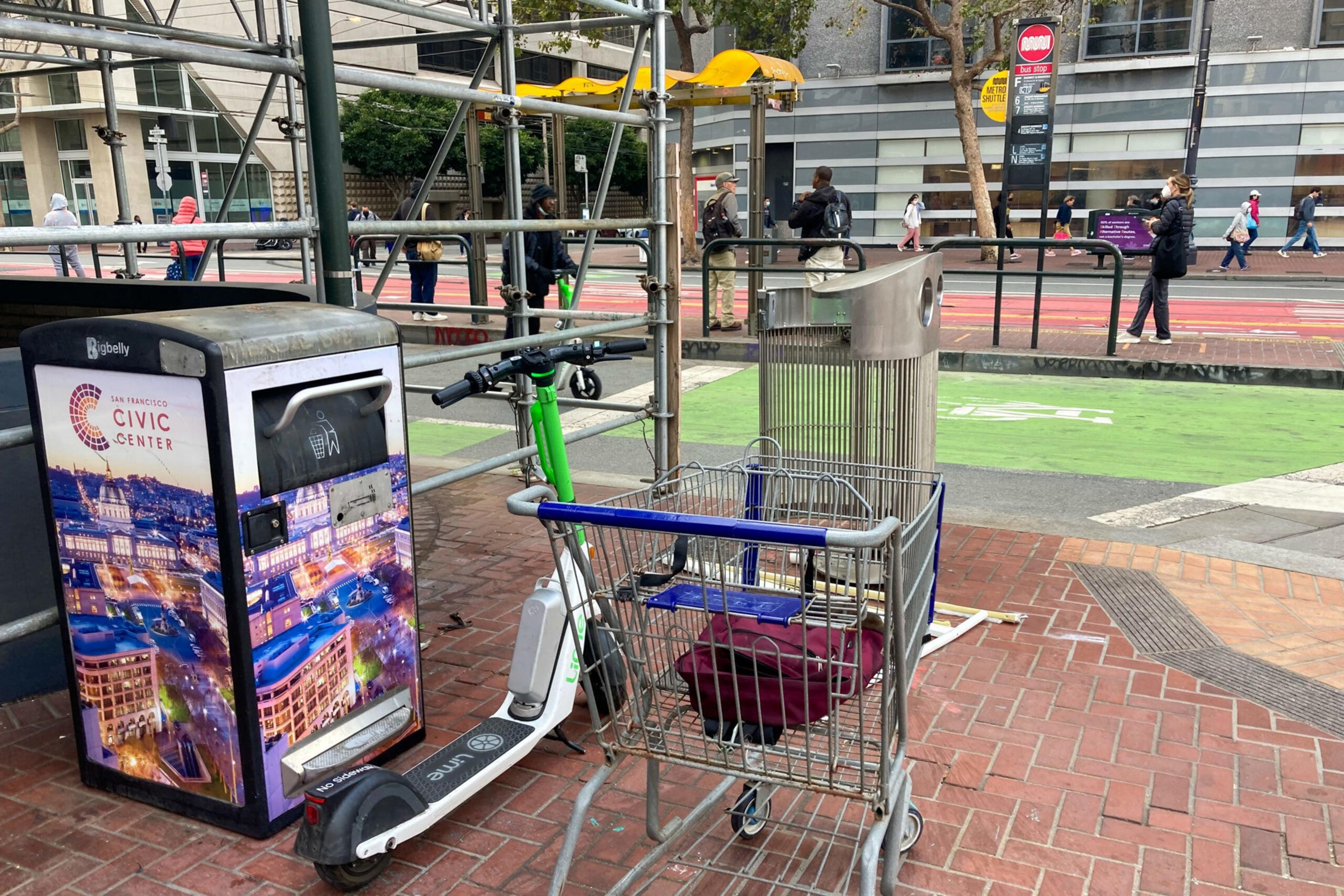 The image shows a public sidewalk with a scooter, a shopping cart, and a trash bin next to scaffolding. People walk nearby, along a street and bike path.