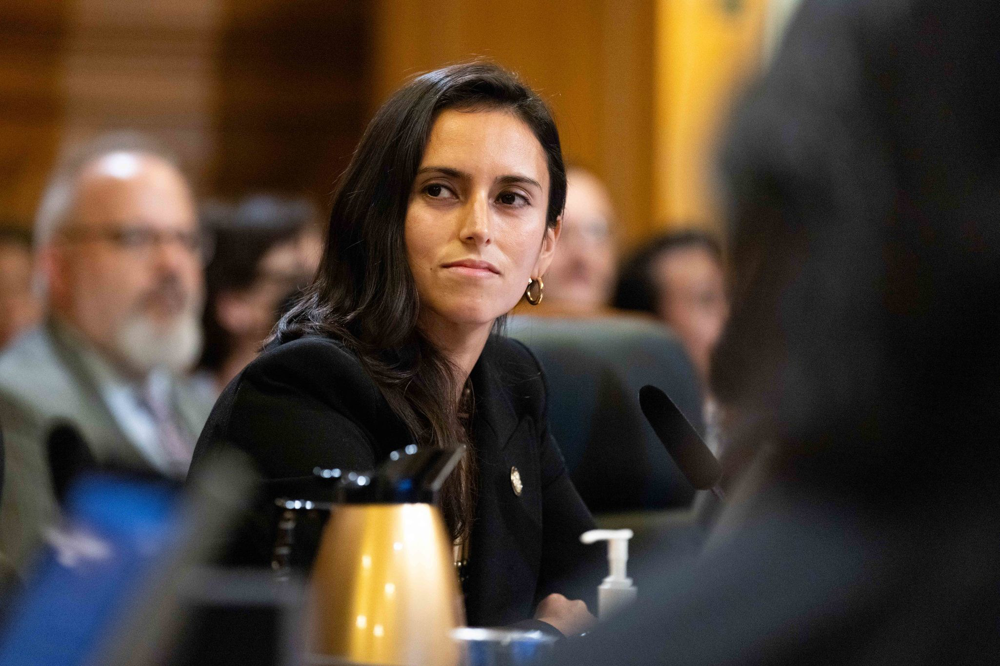 A woman with long dark hair and gold hoop earrings is seated in a formal setting, looking to the side attentively. A gold thermos and sanitizer are visible.