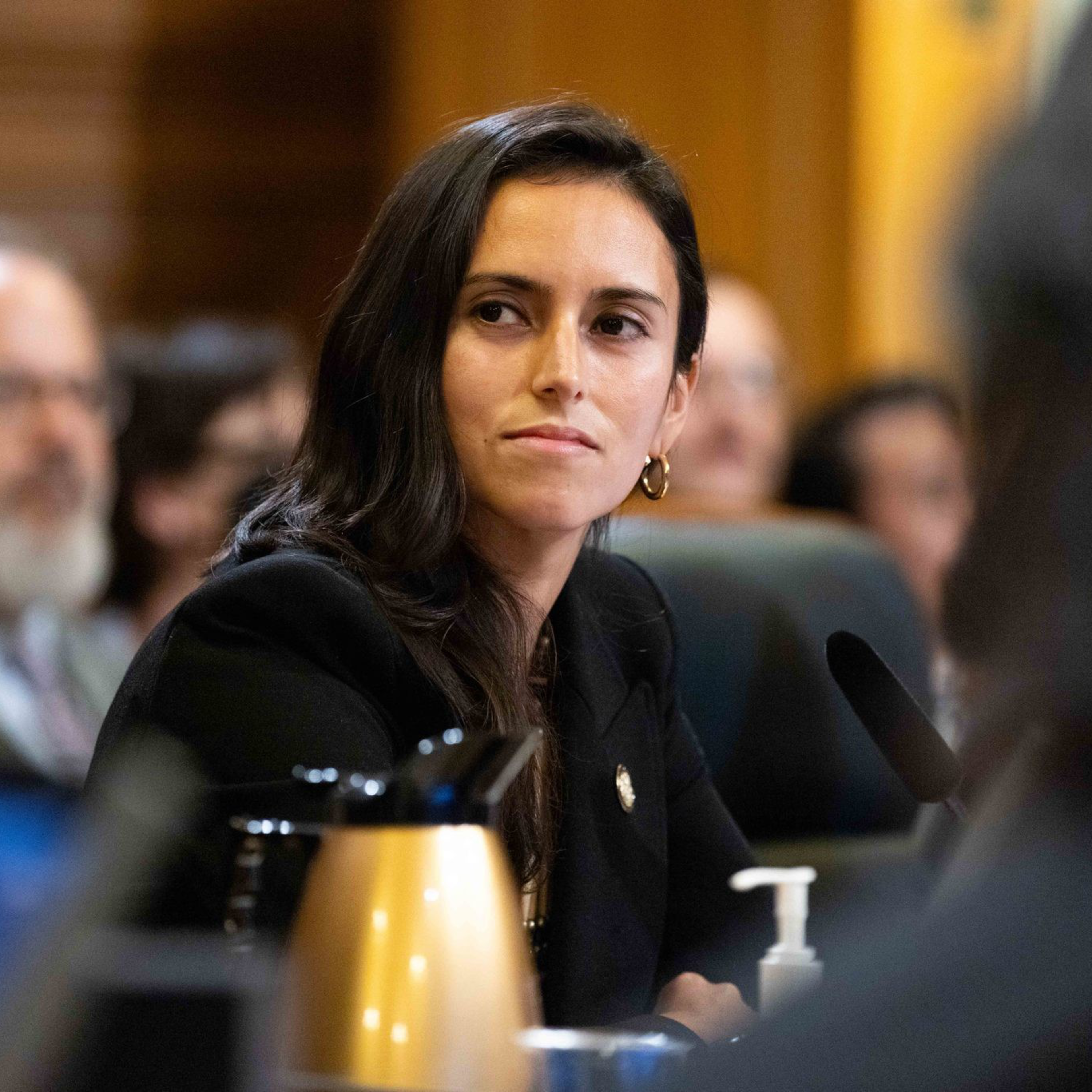 A woman with long dark hair and gold hoop earrings is seated in a formal setting, looking to the side attentively. A gold thermos and sanitizer are visible.