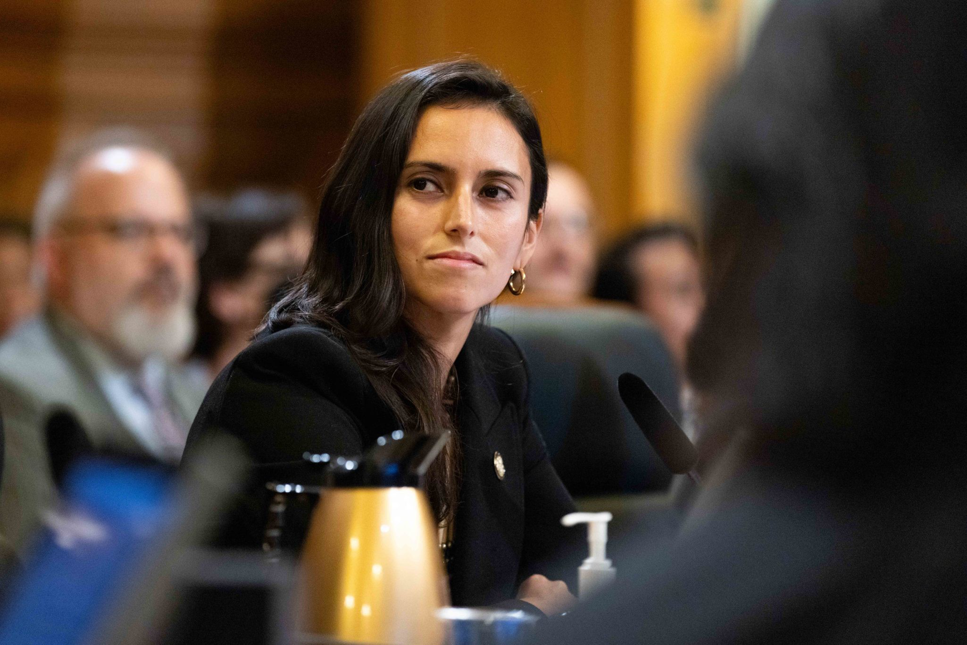A woman with long dark hair and gold hoop earrings is seated in a formal setting, looking to the side attentively. A gold thermos and sanitizer are visible.