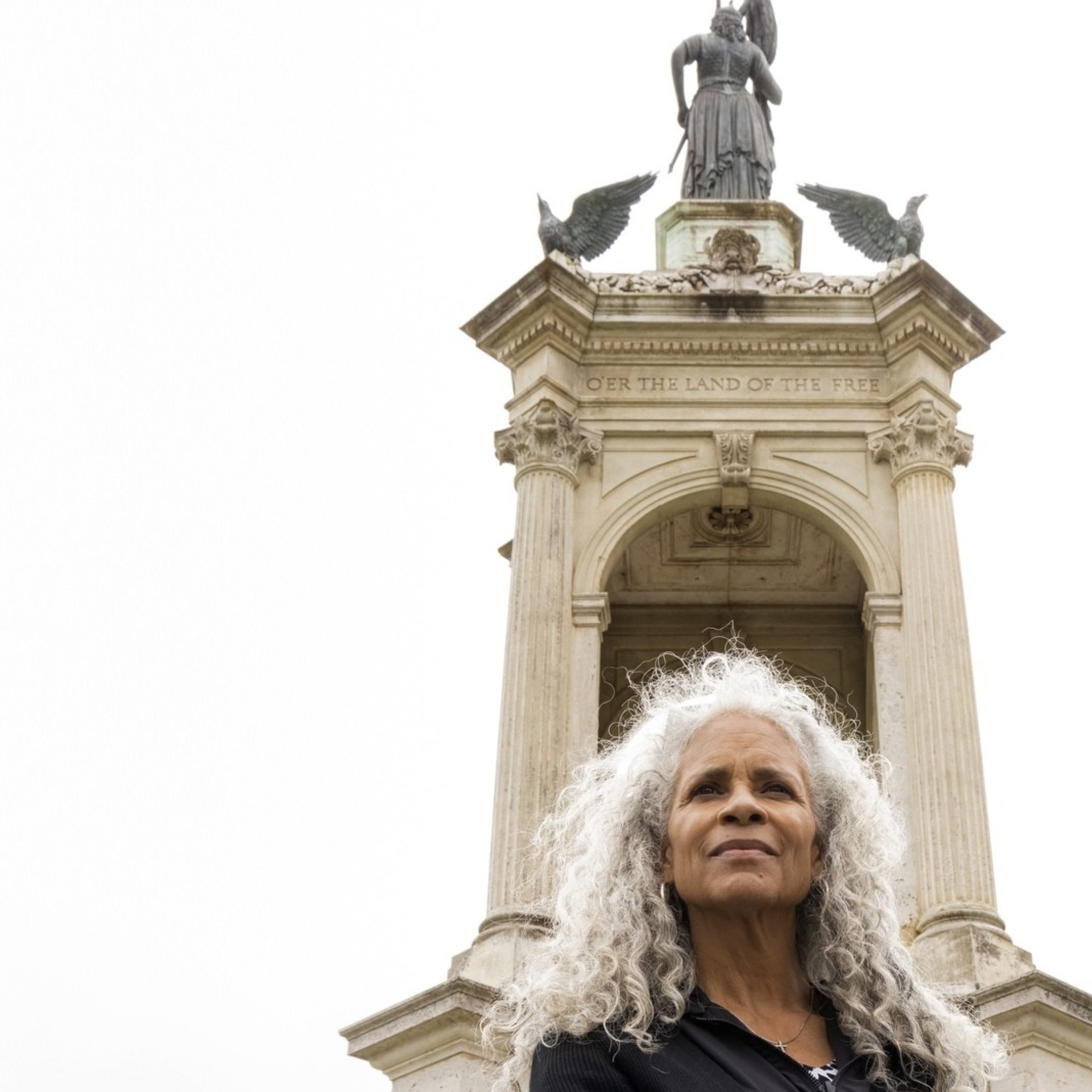 A woman with curly white hair stands confidently with folded arms in front of a tall monument featuring statues and inscriptions against a cloudy sky.