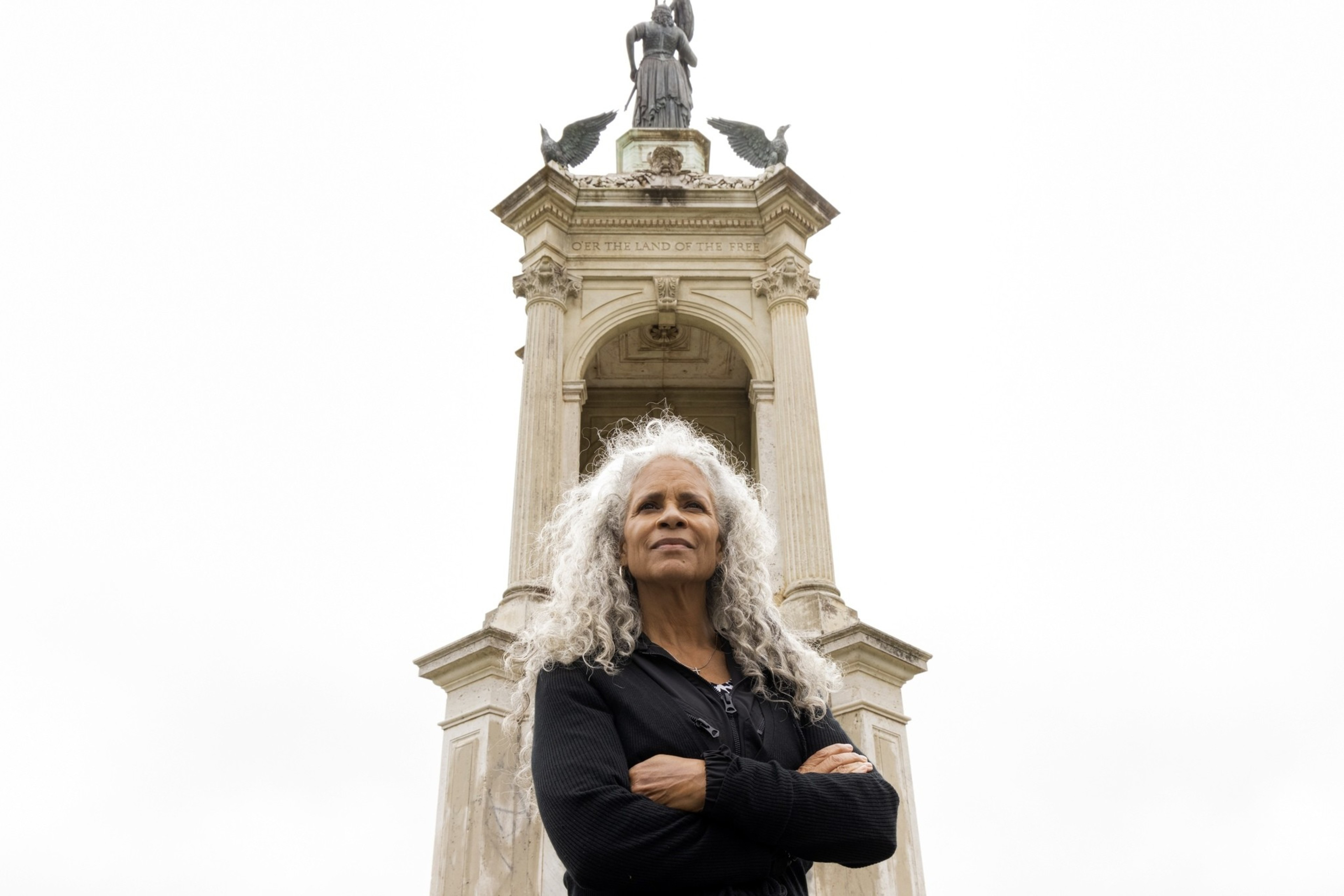 A woman with curly white hair stands confidently with folded arms in front of a tall monument featuring statues and inscriptions against a cloudy sky.