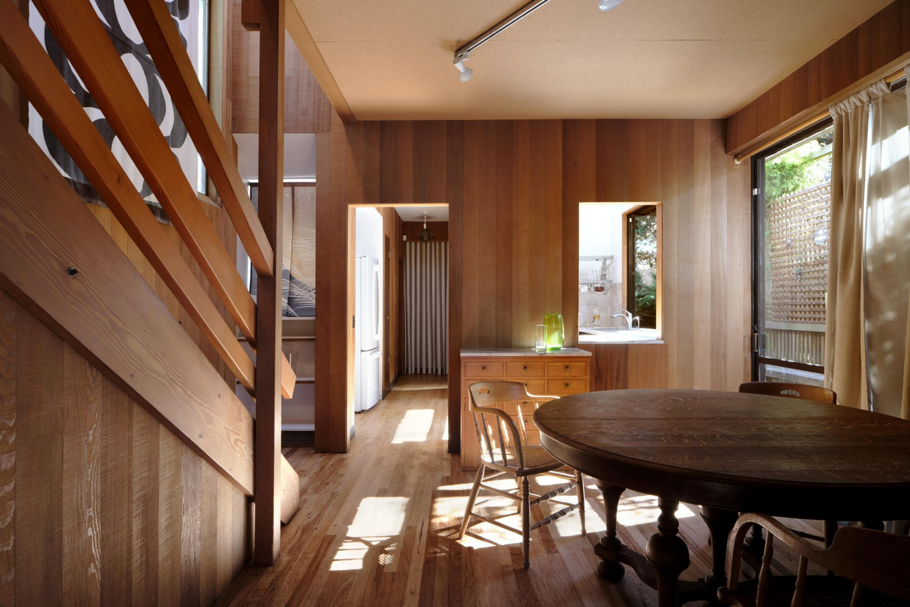 This image shows a cozy wooden interior with a round dining table, chairs, and a staircase. Natural light streams through a window, highlighting warm tones.