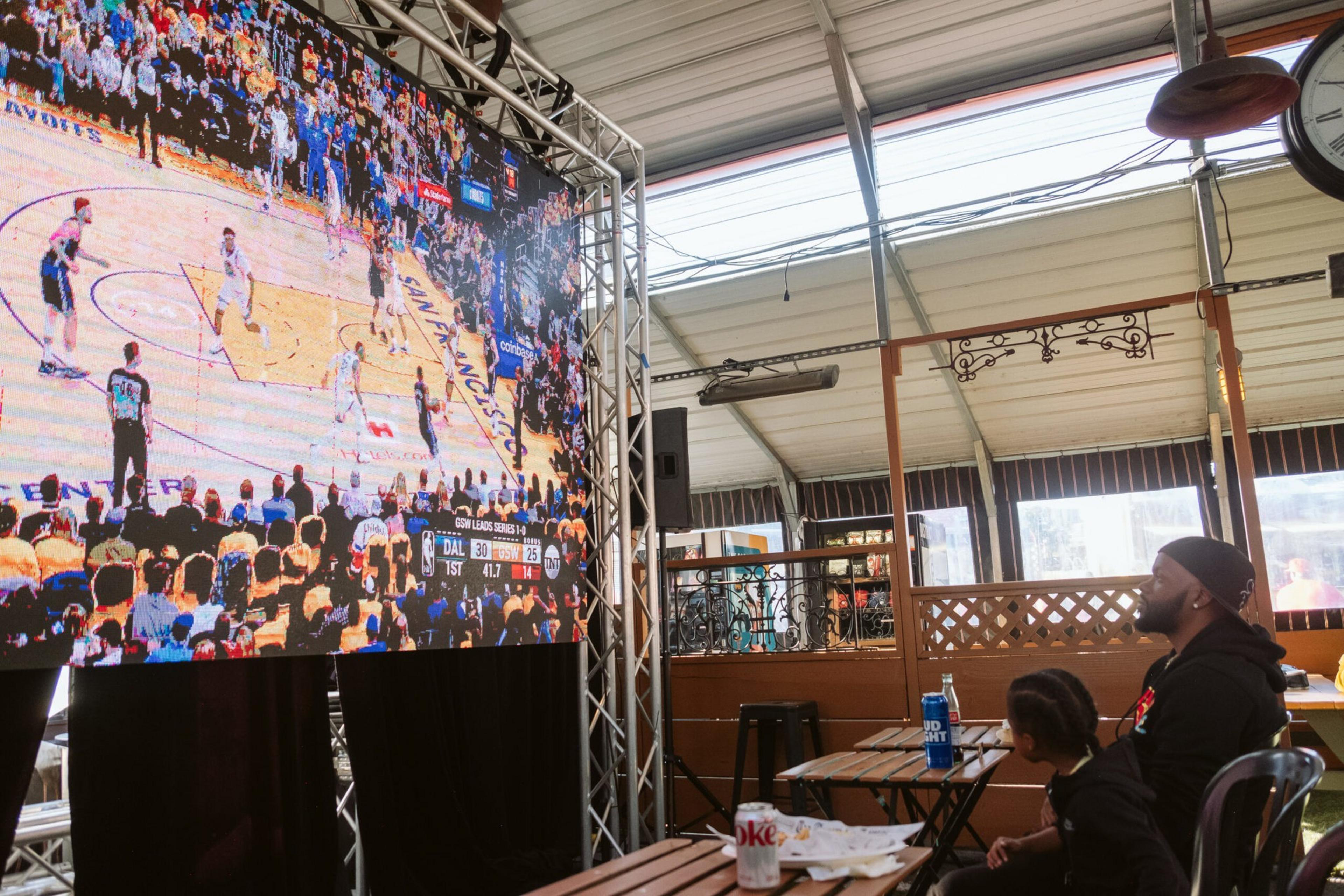 A man and a child sit in a covered outdoor area watching a large screen showing a basketball game between Dallas and Golden State.