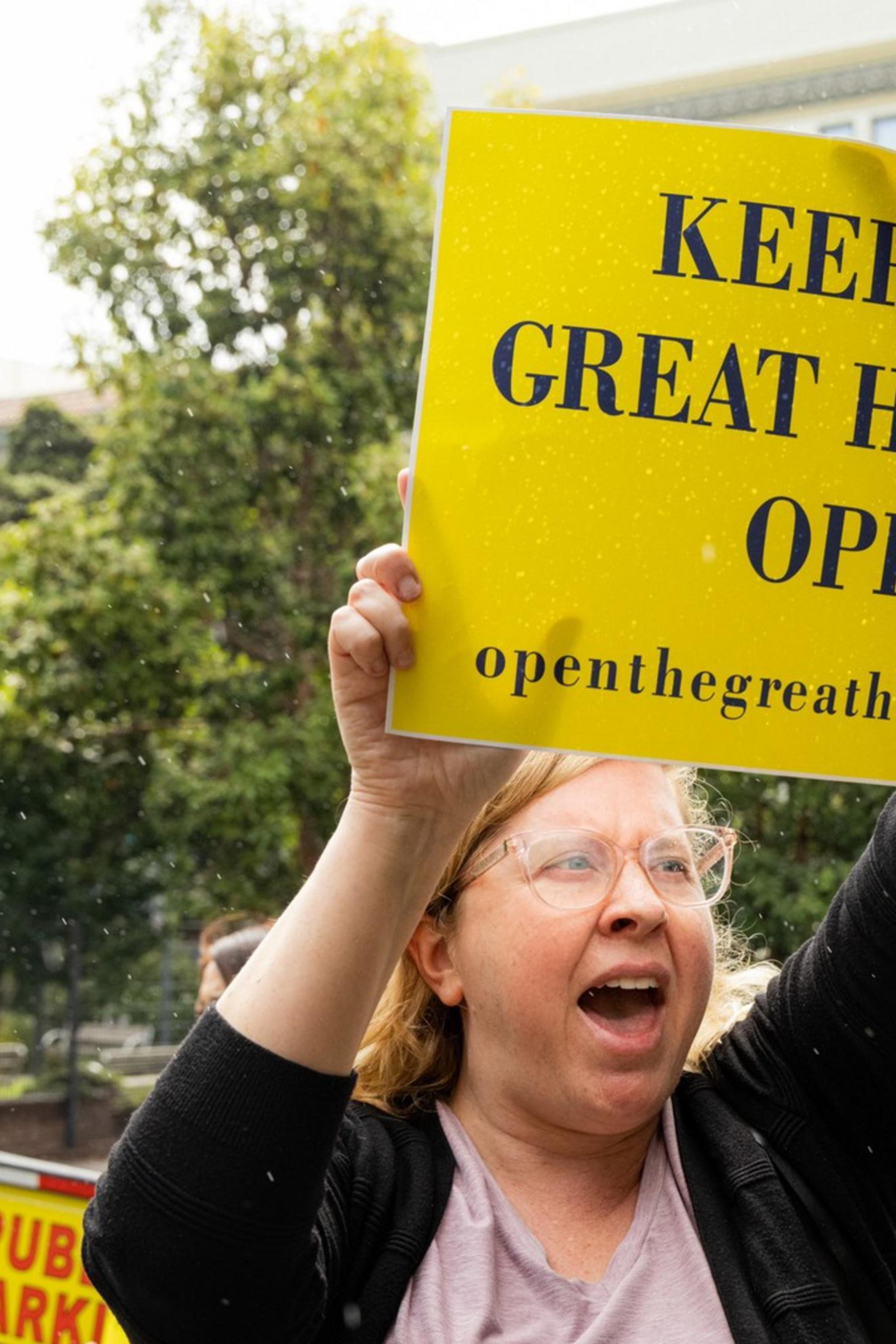 A woman enthusiastically holds a yellow sign reading, &quot;Keep the Great Highway Open&quot; in a city street with trees, buildings, and a school crossing sign visible in the background.