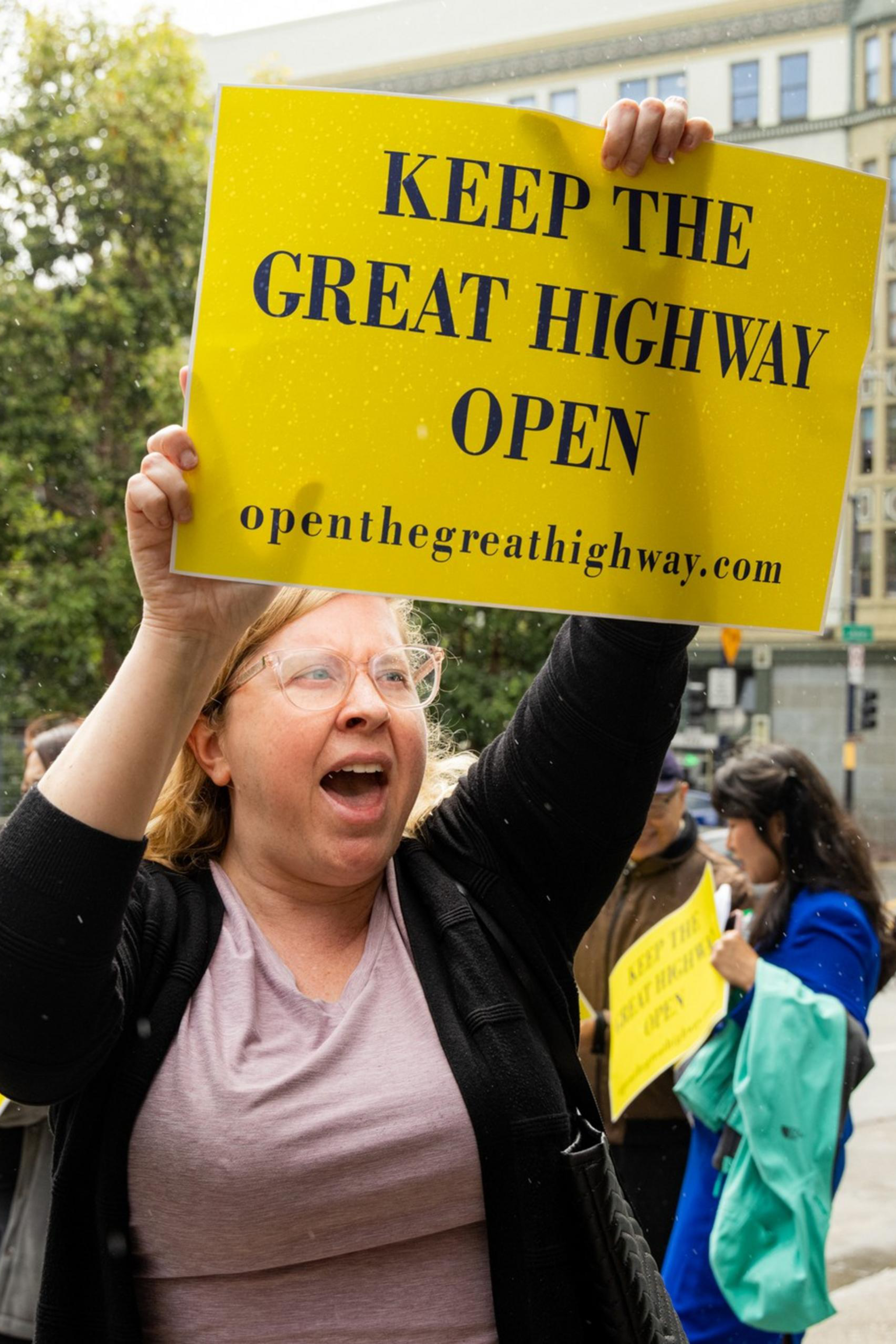 A woman enthusiastically holds a yellow sign reading, &quot;Keep the Great Highway Open&quot; in a city street with trees, buildings, and a school crossing sign visible in the background.