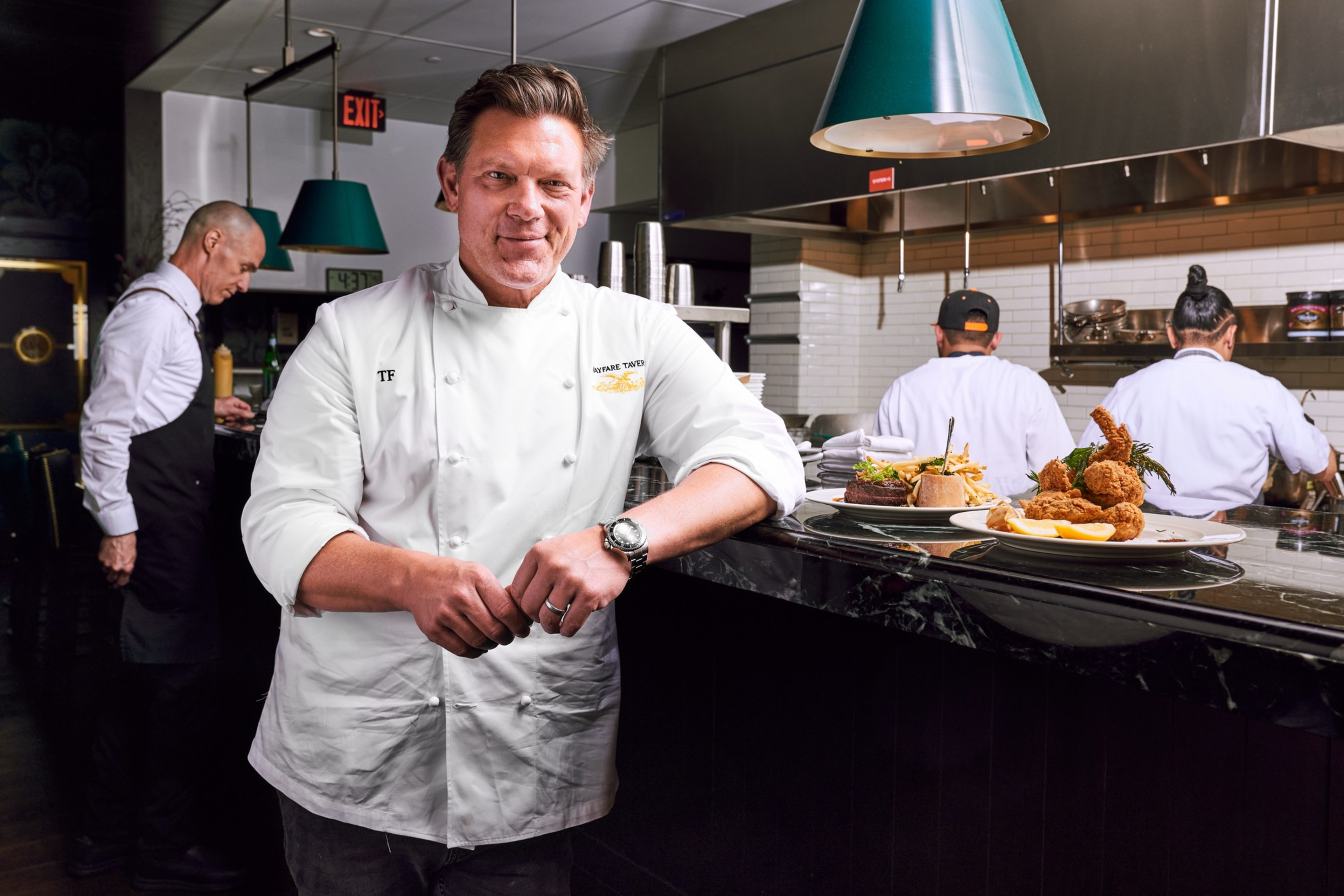 A chef in a white uniform stands confidently in a restaurant kitchen. Behind him, two staff members prepare food. Plates with fried food and sides are on the counter.