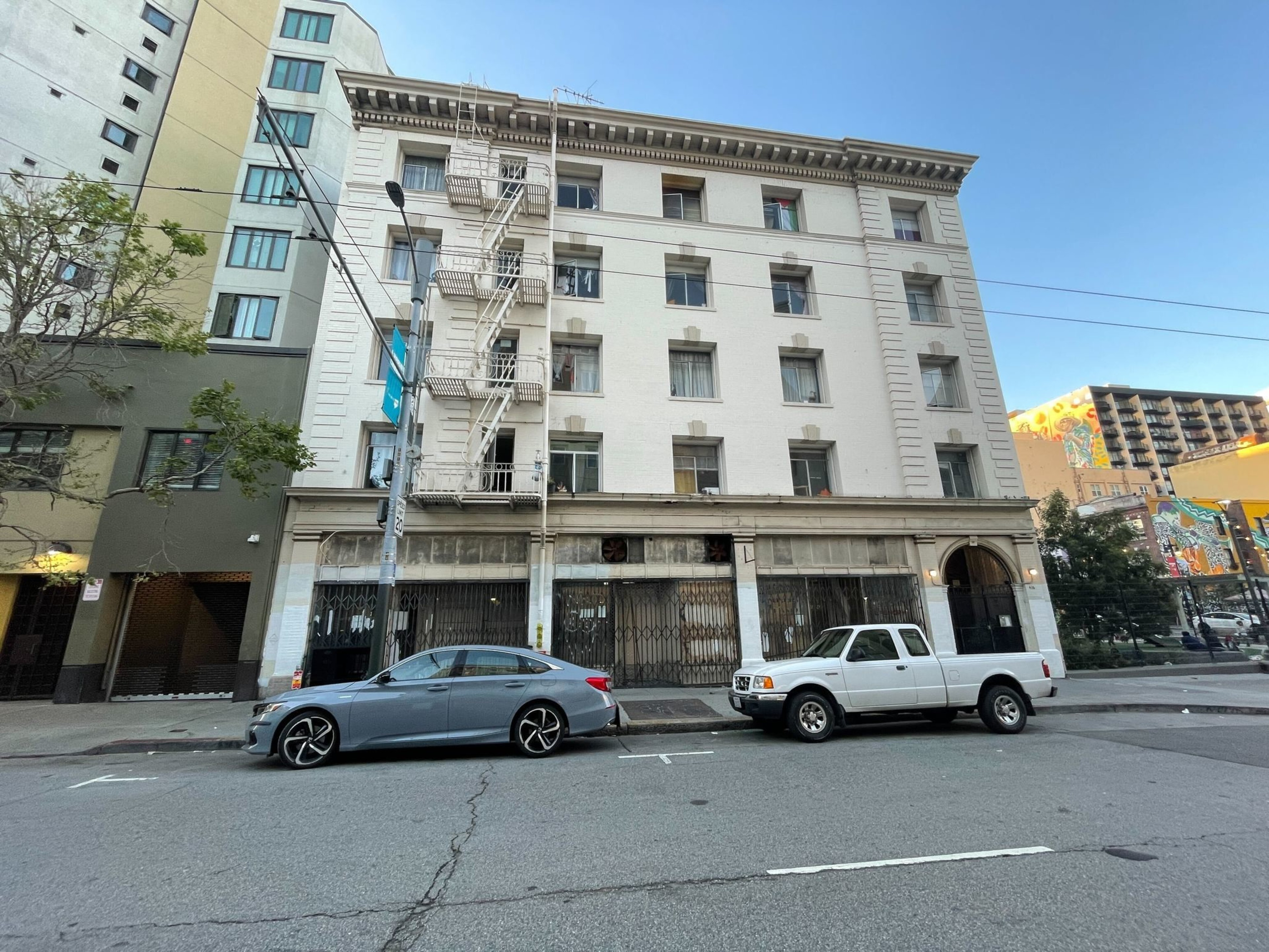 An old building with a fire escape, boarded-up ground floor, parked cars in front, under a clear sky.