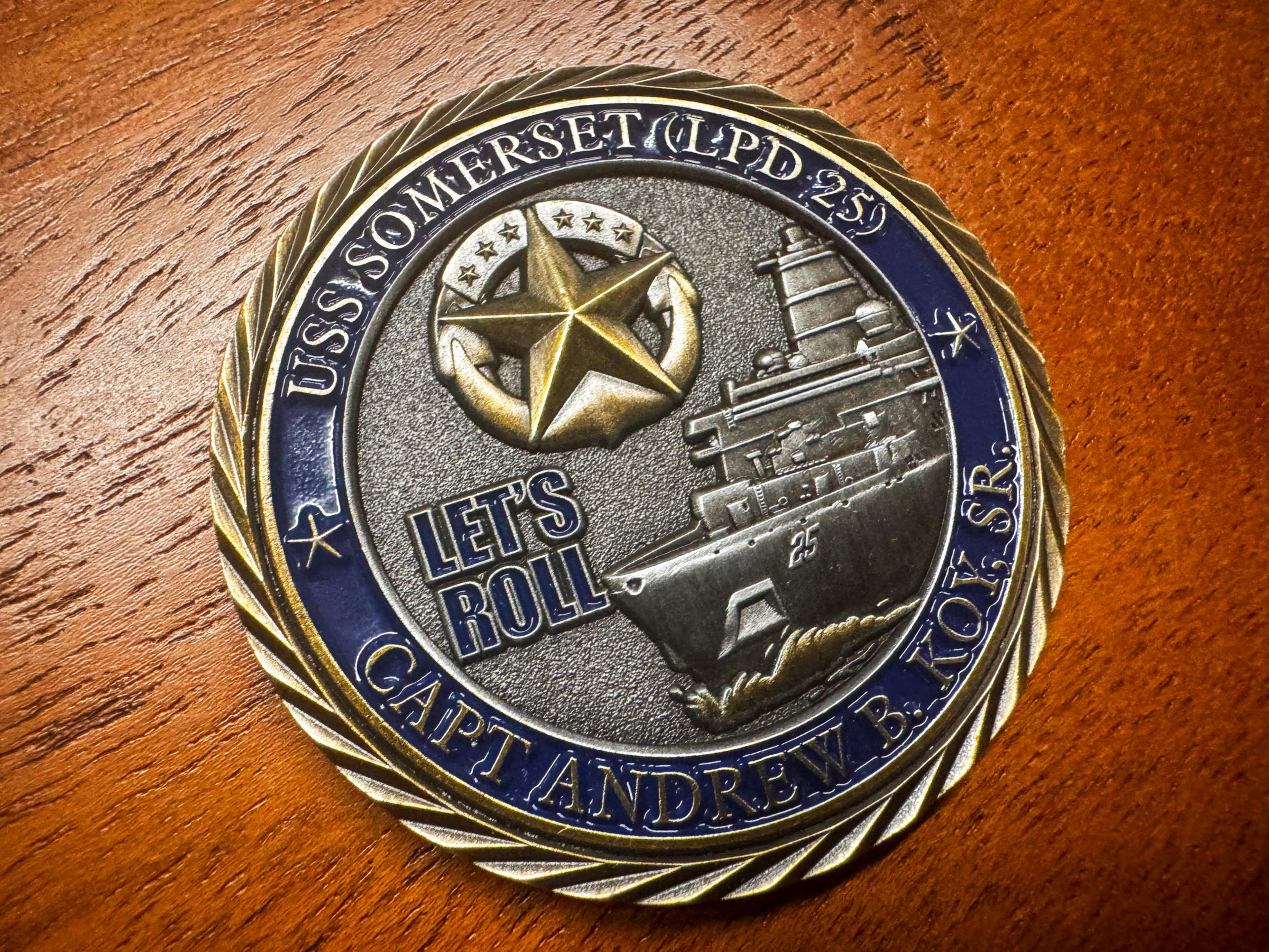 The image shows a round commemorative coin with "USS Somerset (LPD 25)" and "Let's Roll" engraved, featuring a ship and a gold star on a wooden surface.