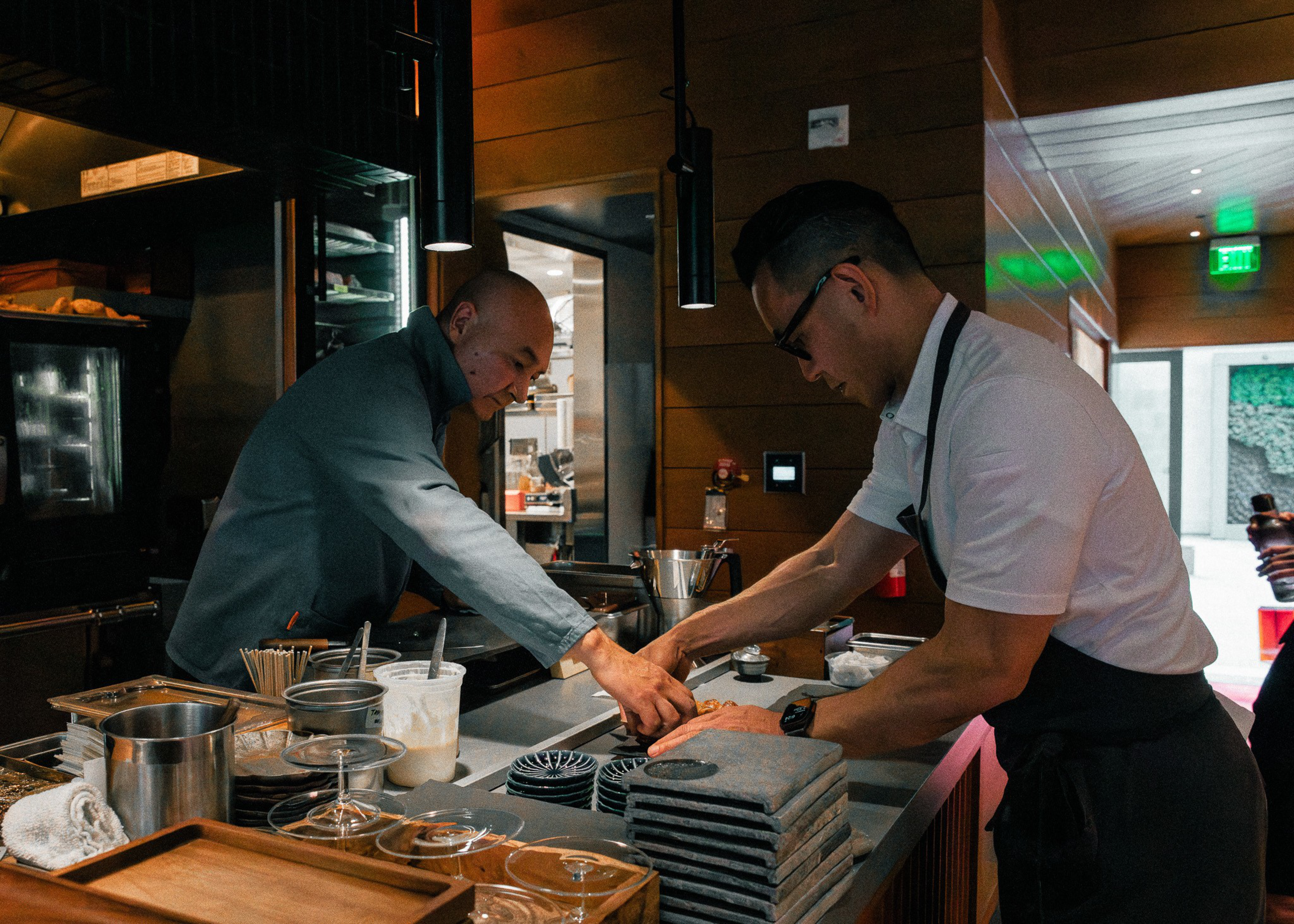 Two men, one in a blue jacket and the other in a white shirt and apron, shake hands over a kitchen counter with utensils and stacked trays.