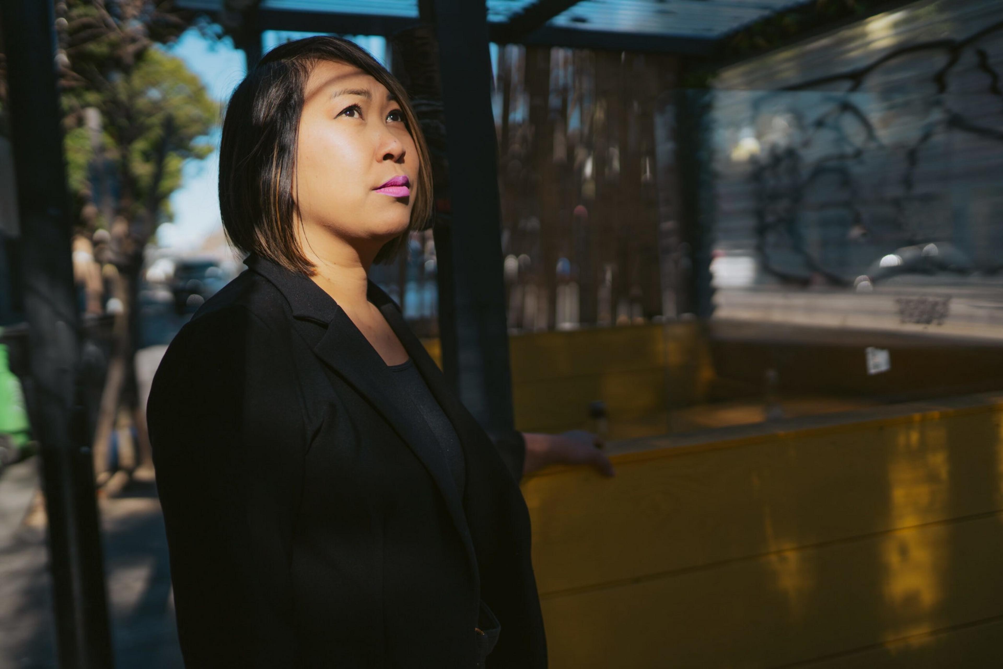 A woman with short hair and bright pink lipstick stands in partial sunlight, wearing a black jacket, looking thoughtfully upwards outdoors.