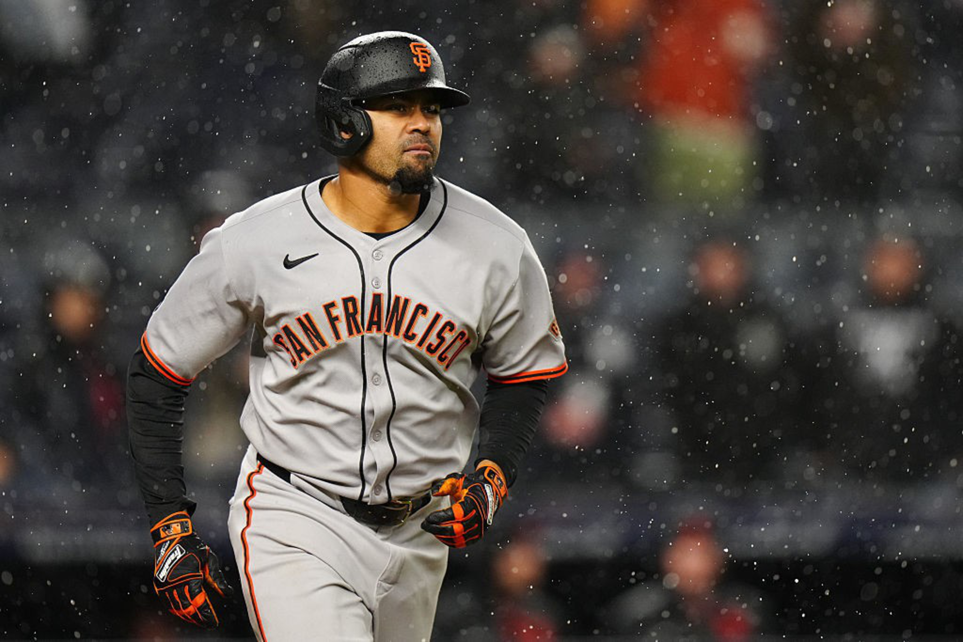 A baseball player in a gray &quot;San Francisco&quot; uniform runs during a game in the rain. He wears a helmet and black gloves, with blurred spectators in the background.