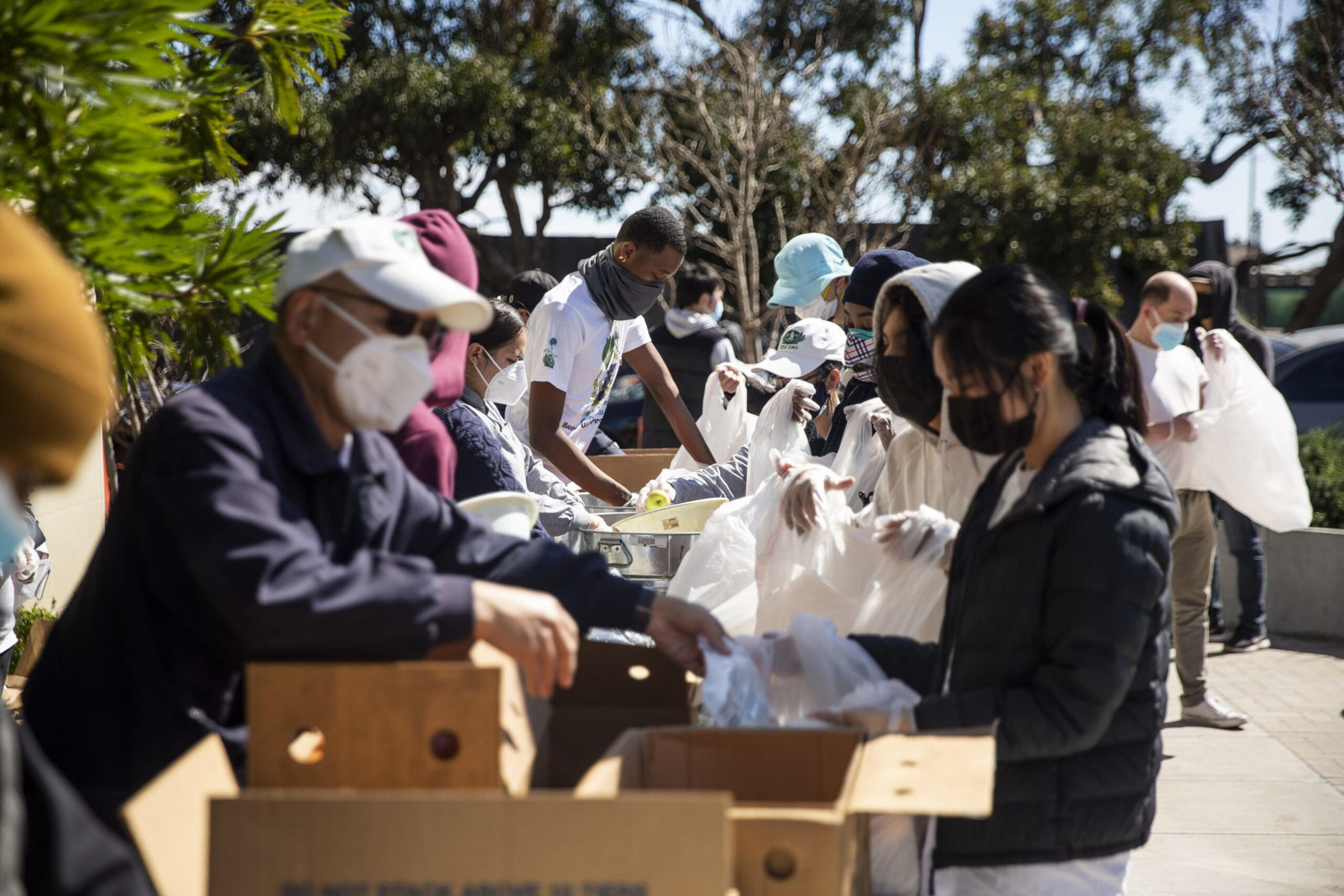 People wearing masks and gloves pack food into plastic bags outdoors, standing in a line near cardboard boxes and trees.