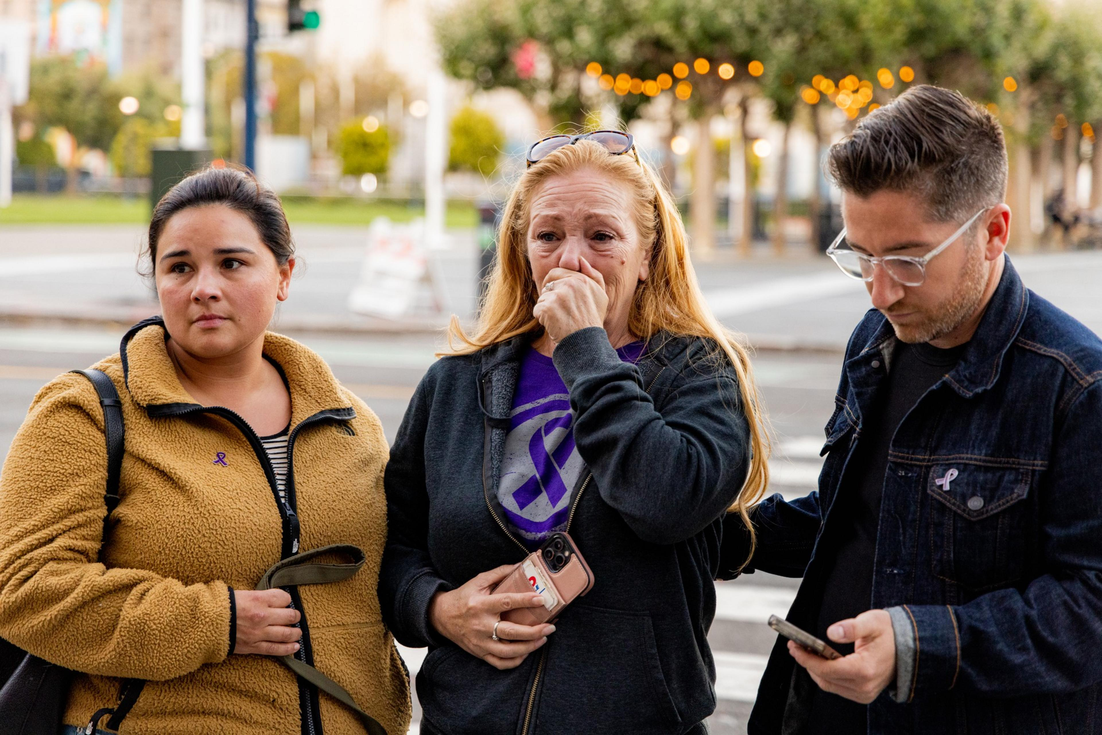 Three people stand outdoors; a woman wipes tears, clutching a phone, flanked by a serious-looking woman and a man looking down at his phone.