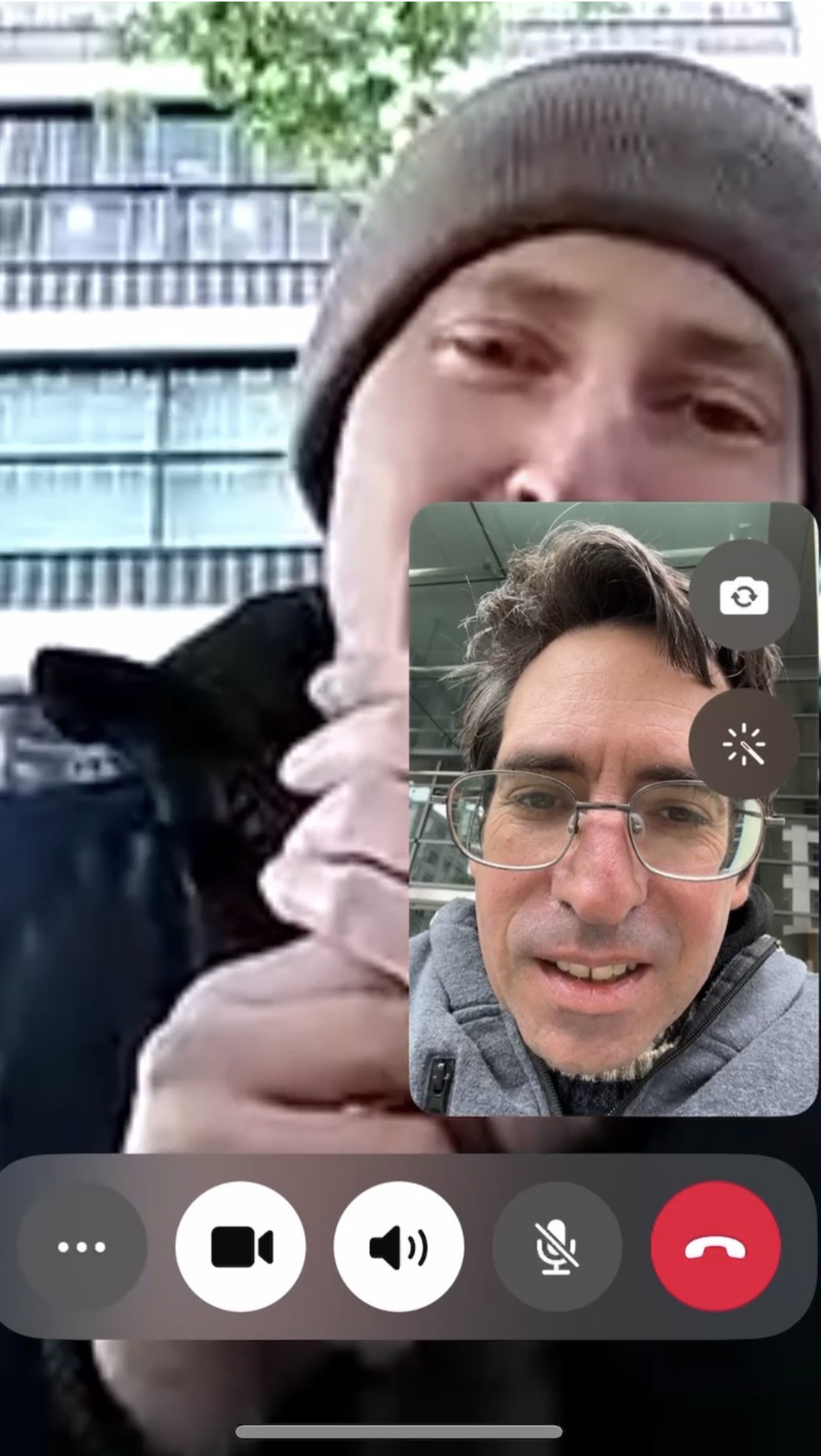 A man in a gray beanie and dark jacket is outdoors near a building, holding something close to his mouth, while another man with glasses and messy hair appears in a video call inset.