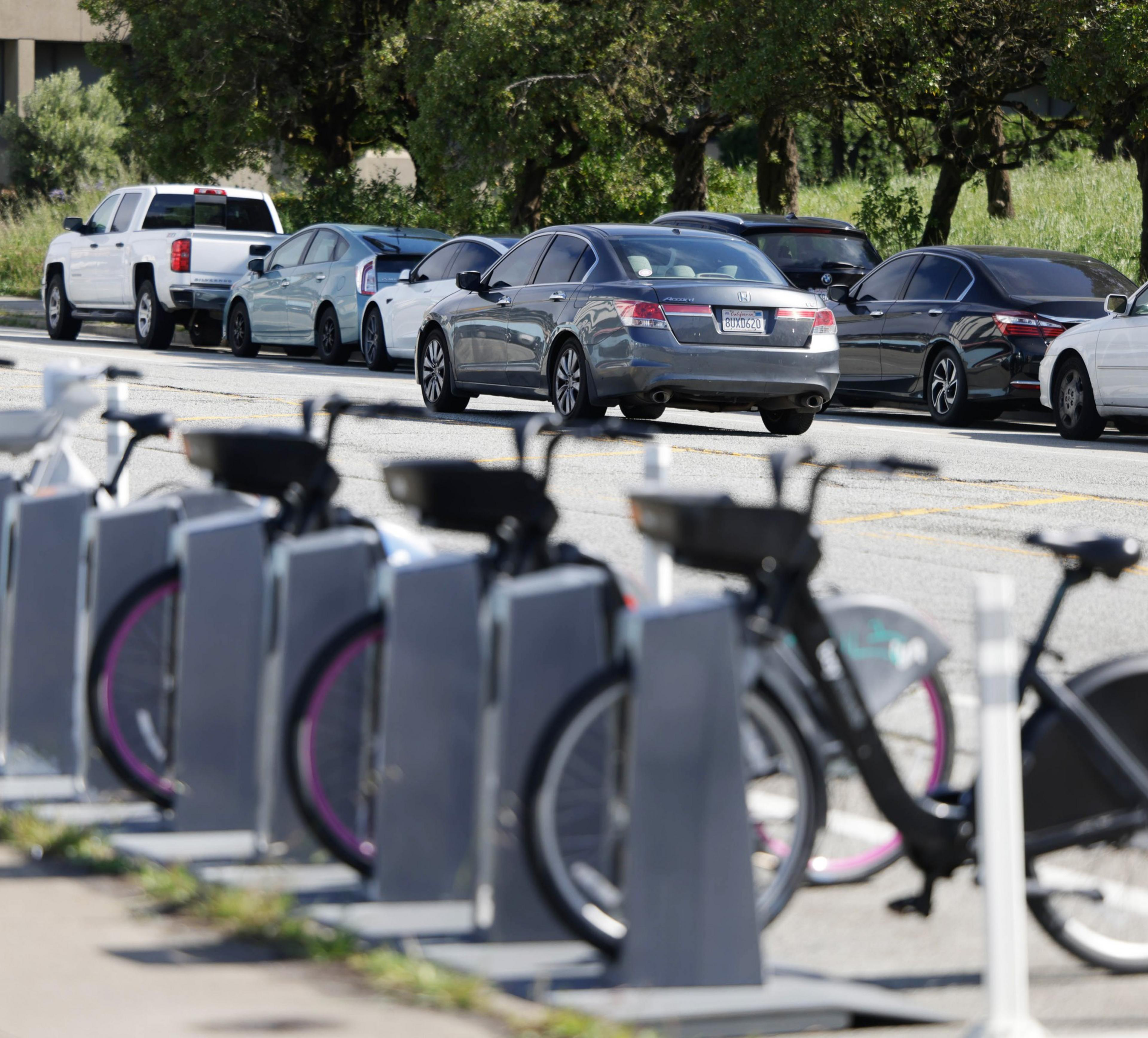 A row of docked rental bikes in the foreground with a line of cars on a road behind them.