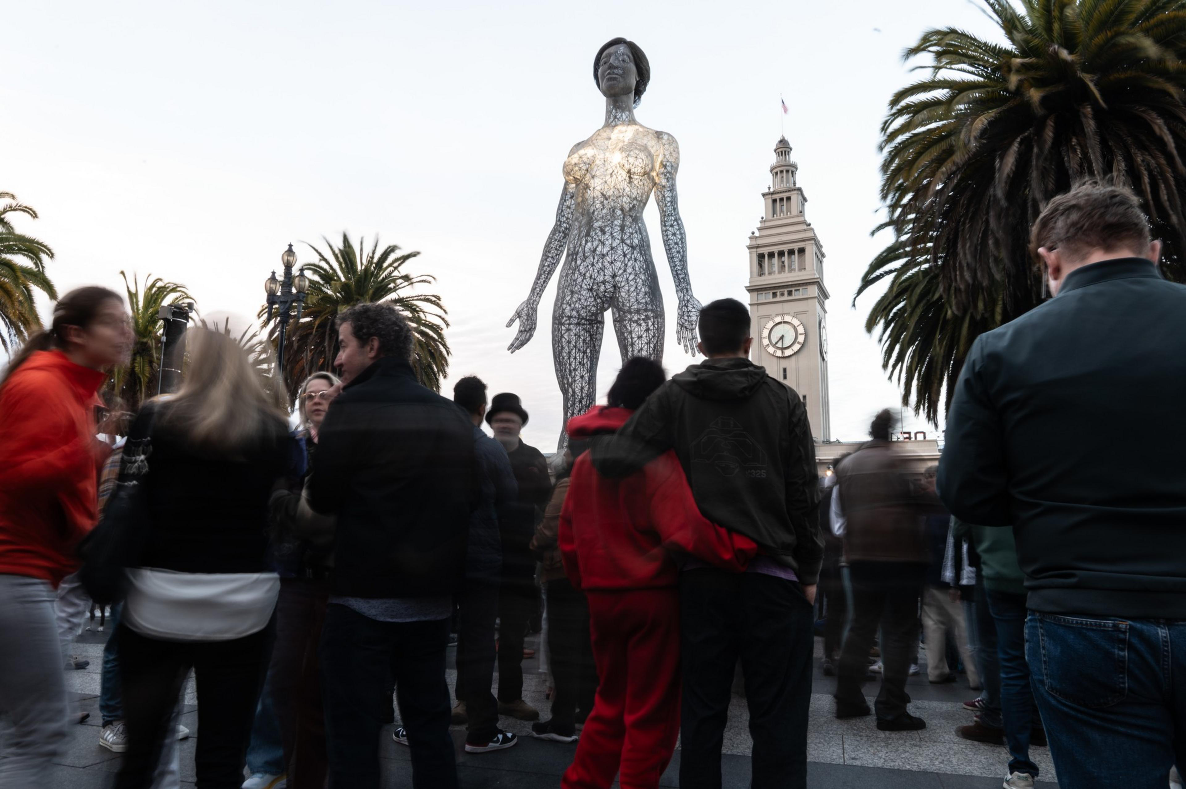 A crowd gathers around a large, illuminated sculpture of a figure with a lattice structure, set against palm trees and a historic clock tower.