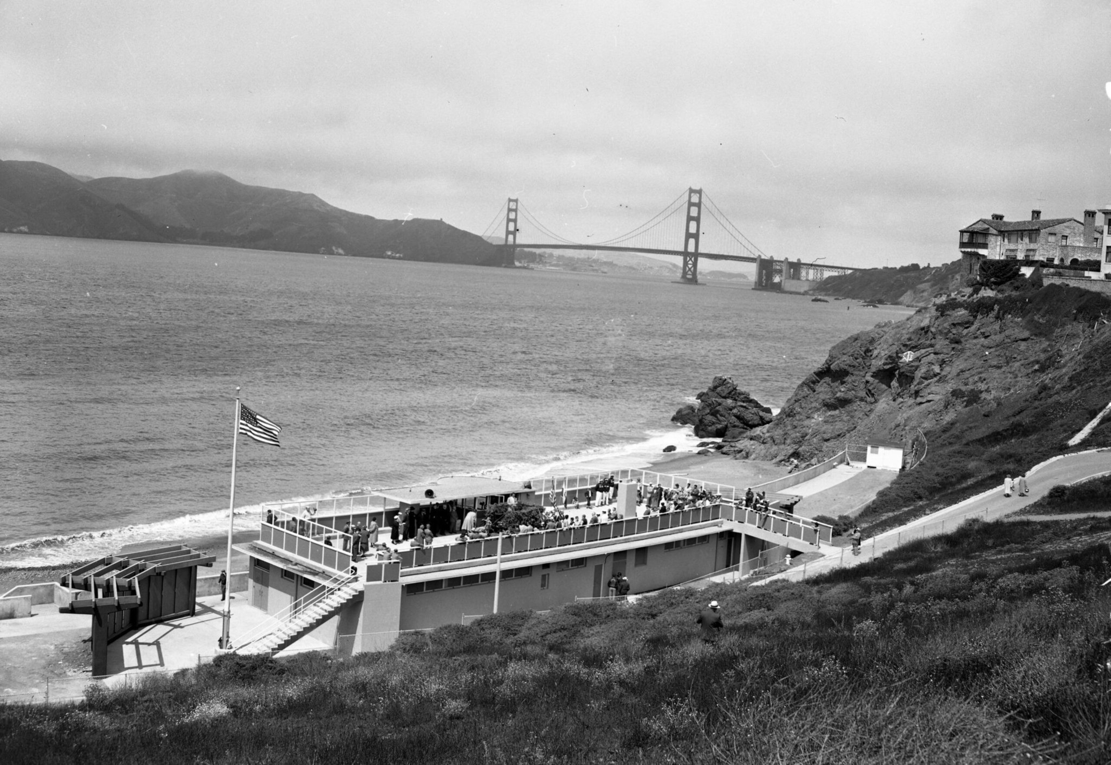 A group of people gather on a flat-roofed building by the shore with the Golden Gate Bridge and hills in the background.