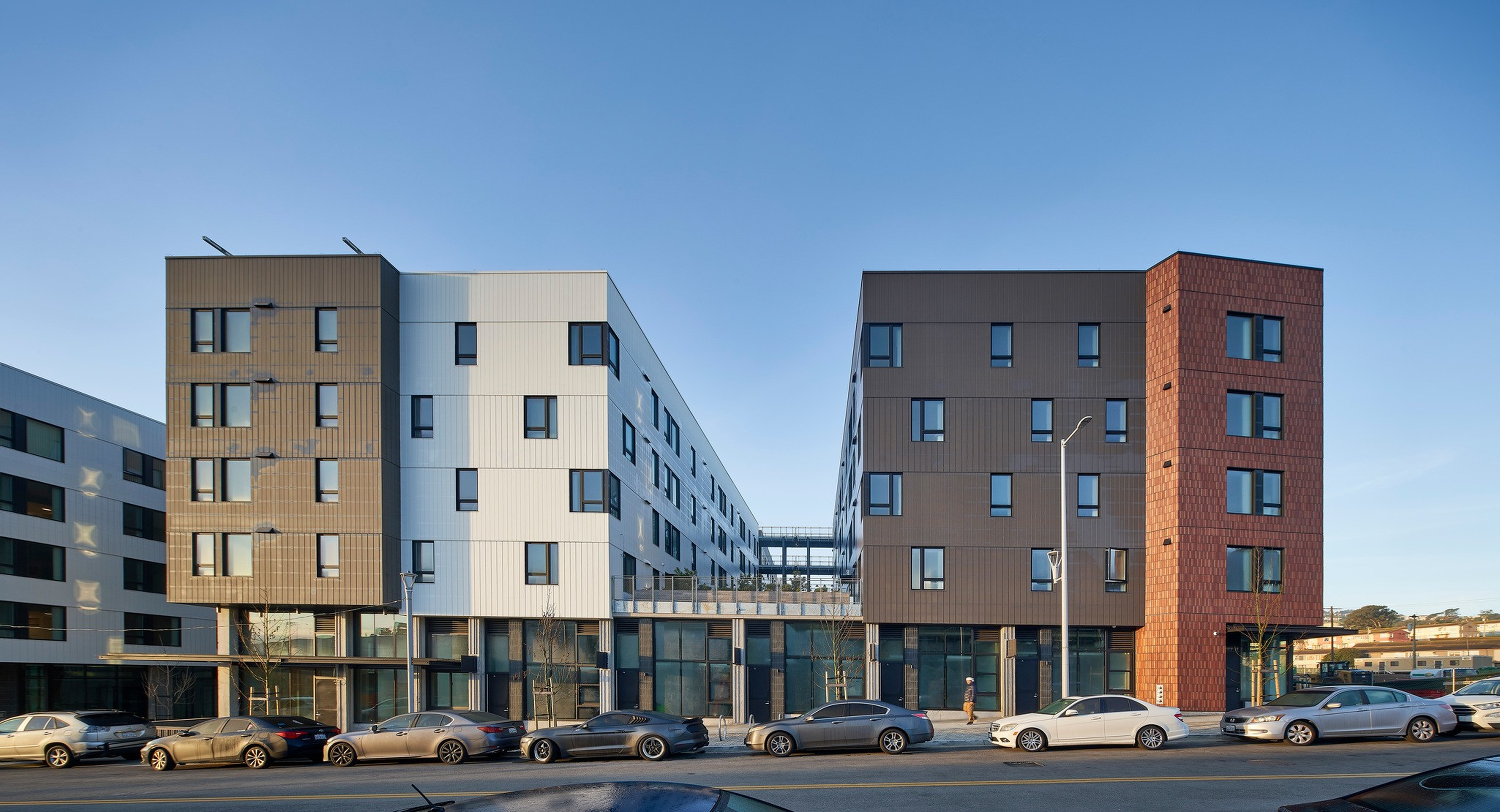 Two modern apartment buildings with different colored panels, tan and white on the left, brown and rust on the right, are separated by a walkway with parked cars in front.