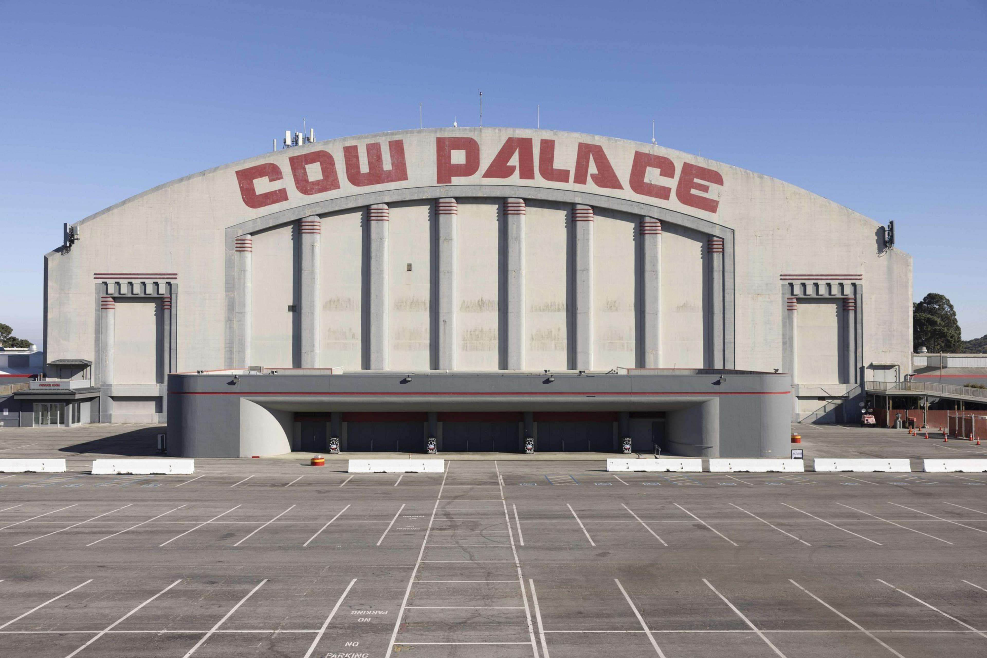A large, grey building with &quot;COW PALACE&quot; in red letters, featuring an empty parking lot and a clear, blue sky above.