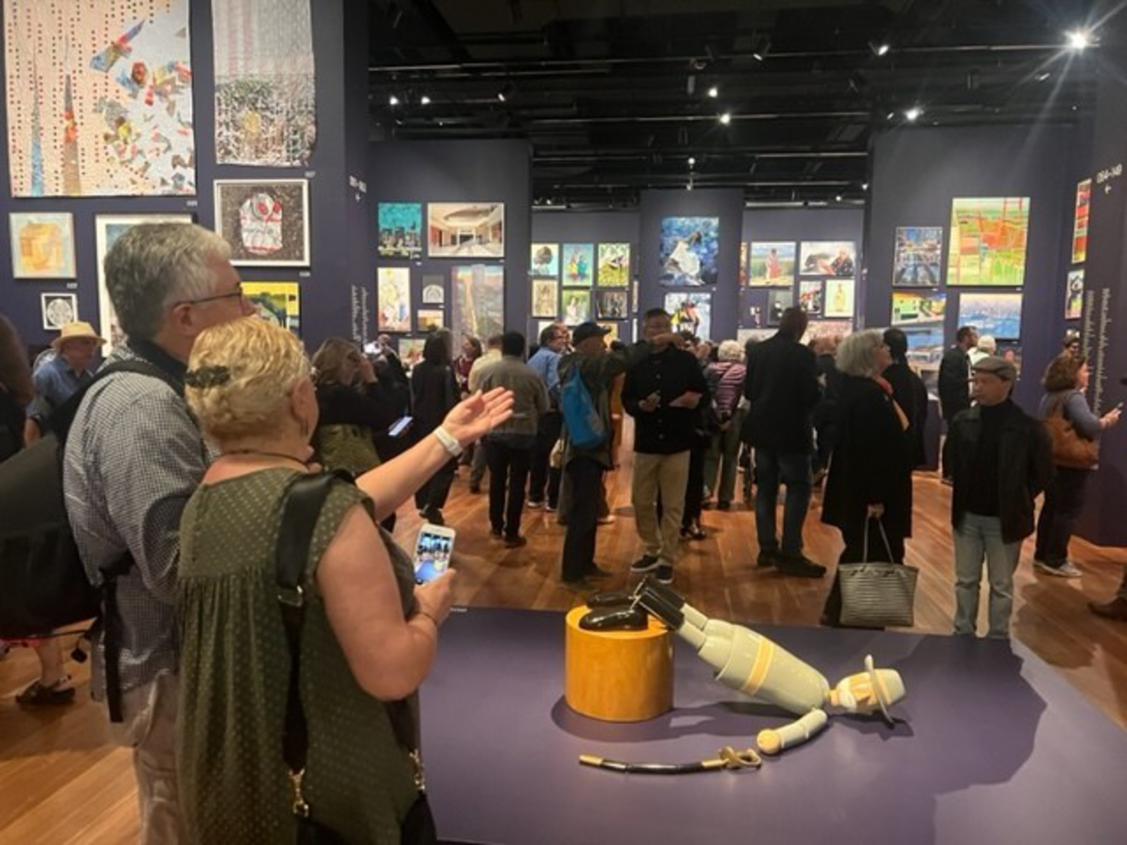 People view and discuss various paintings in a crowded art gallery, with a wooden puppet figure lying on the floor in the foreground.