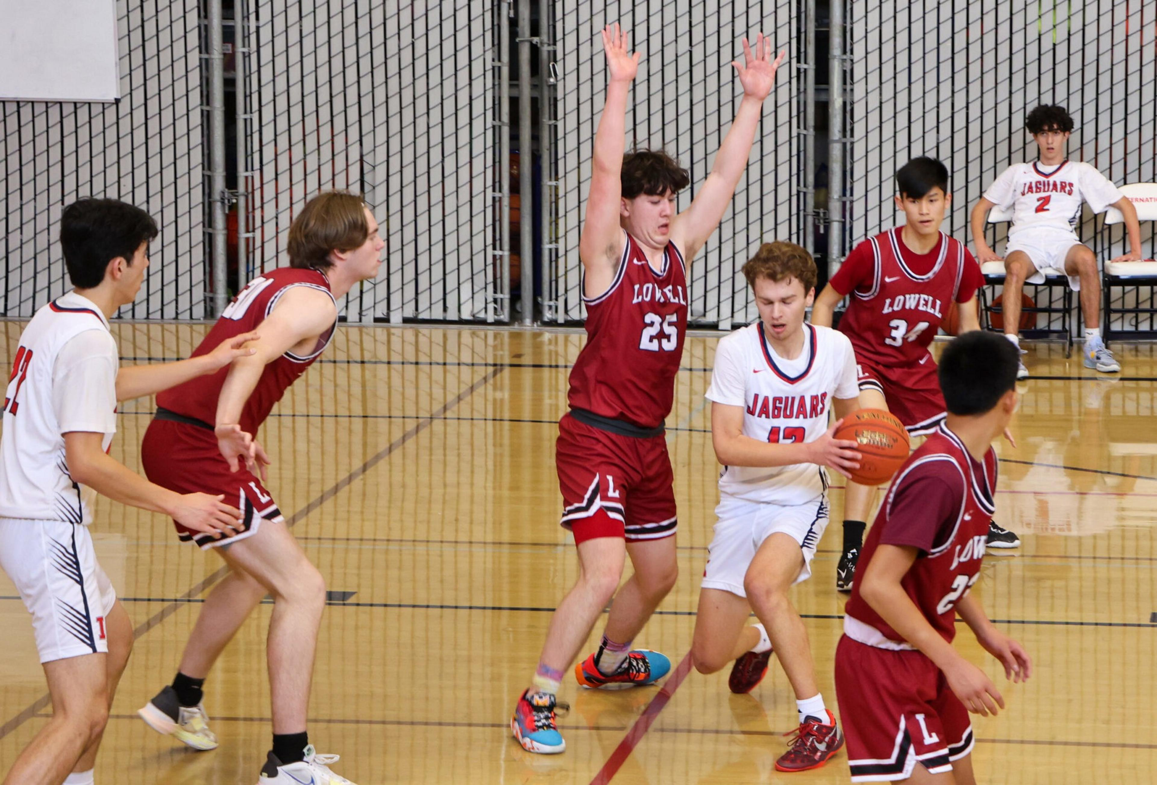 A basketball player in white dribbles while four defenders in maroon raise arms and position themselves; one player in white sits on the bench.