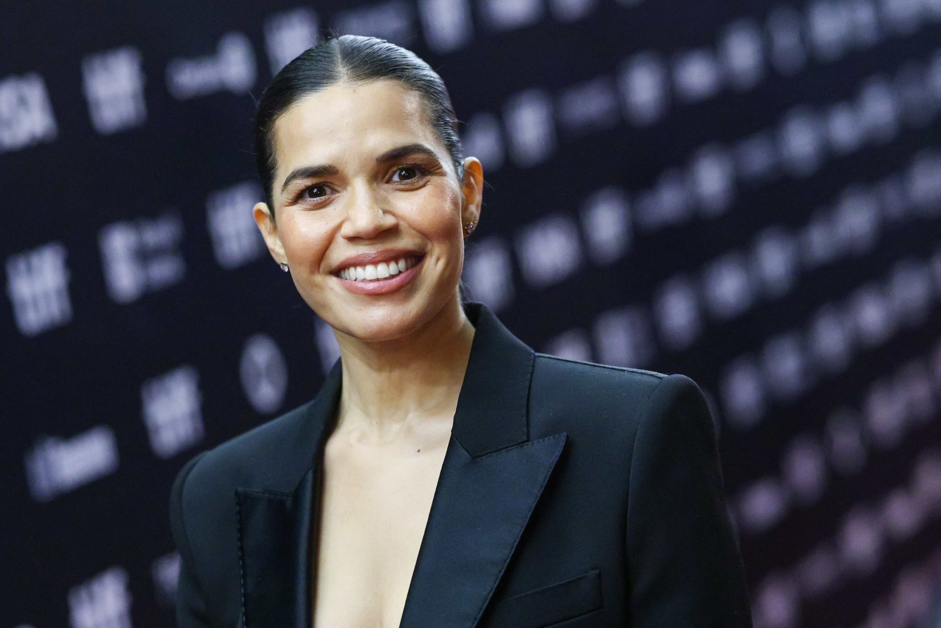 A woman with dark hair tied back smiles warmly, wearing a black blazer, standing against a blurred dark backdrop with light text.