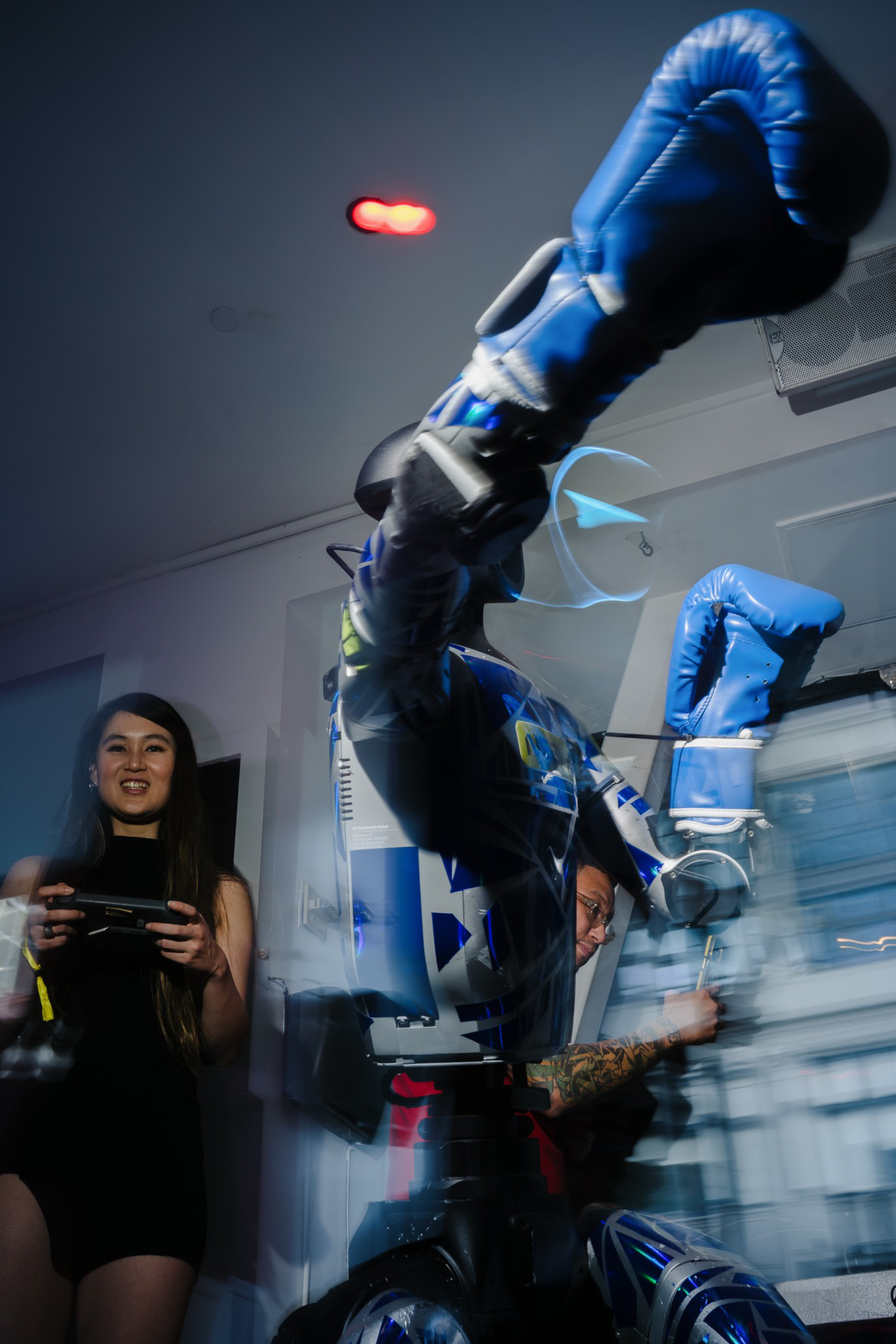 A person in a black dress controls a boxing robot with blue gloves, while a tattooed man adjusts the robot’s arm in a room with blurred motion.