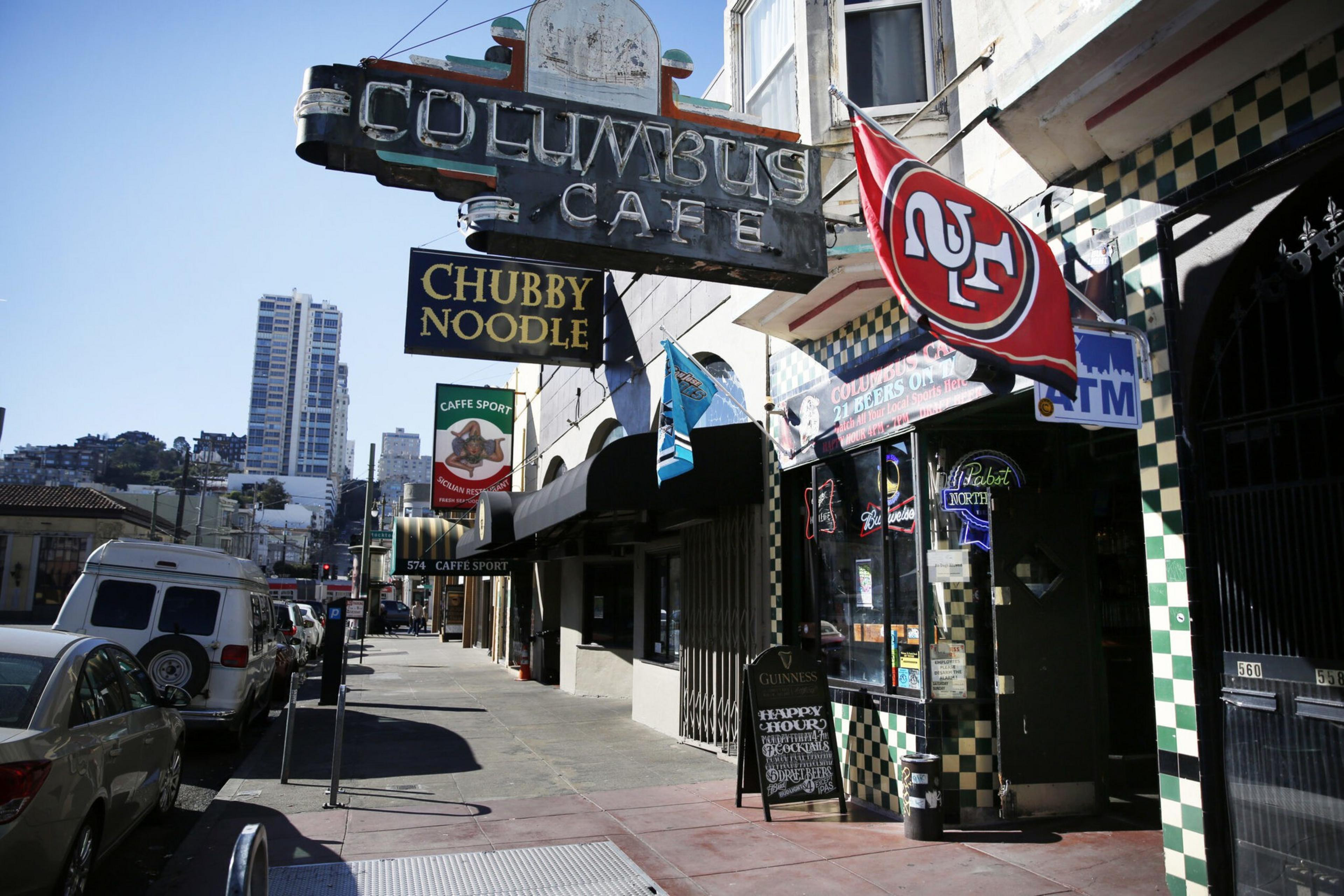 A street view with signs for Columbus Cafe, Chubby Noodle, and Caffe Sport, featuring a 49ers flag and parked cars along the sidewalk.