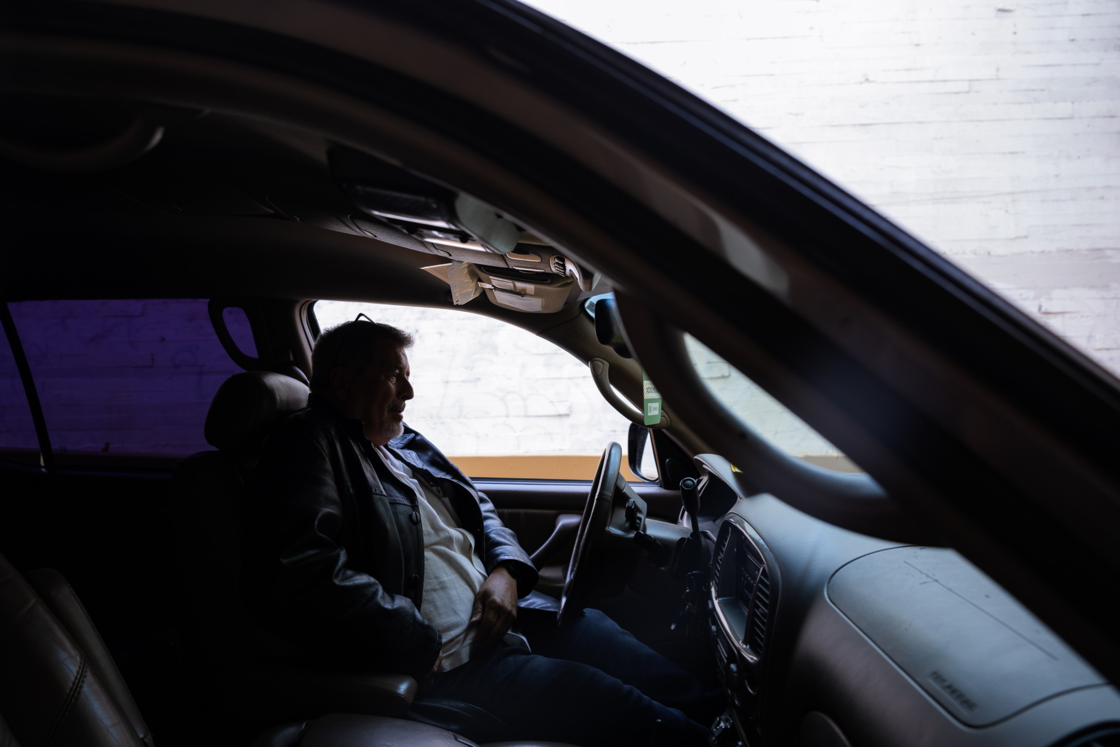 A man wearing a leather jacket sits in the driver’s seat of a parked car, facing forward, with a white brick wall visible outside.