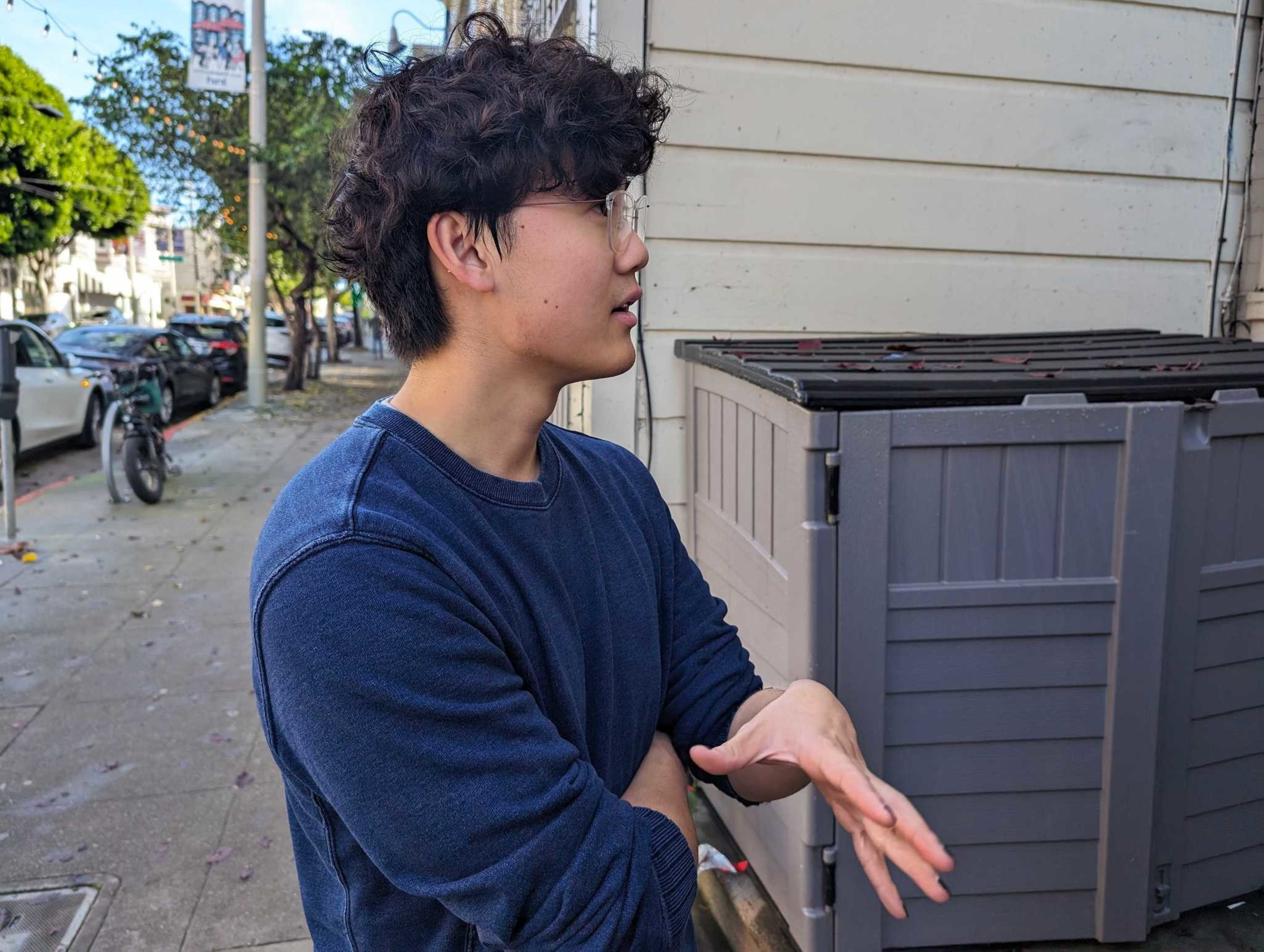 A young man with curly brown hair and clear horn-rimmed glasses and wearing a blue sweater stands on a city sidewalk and talks.